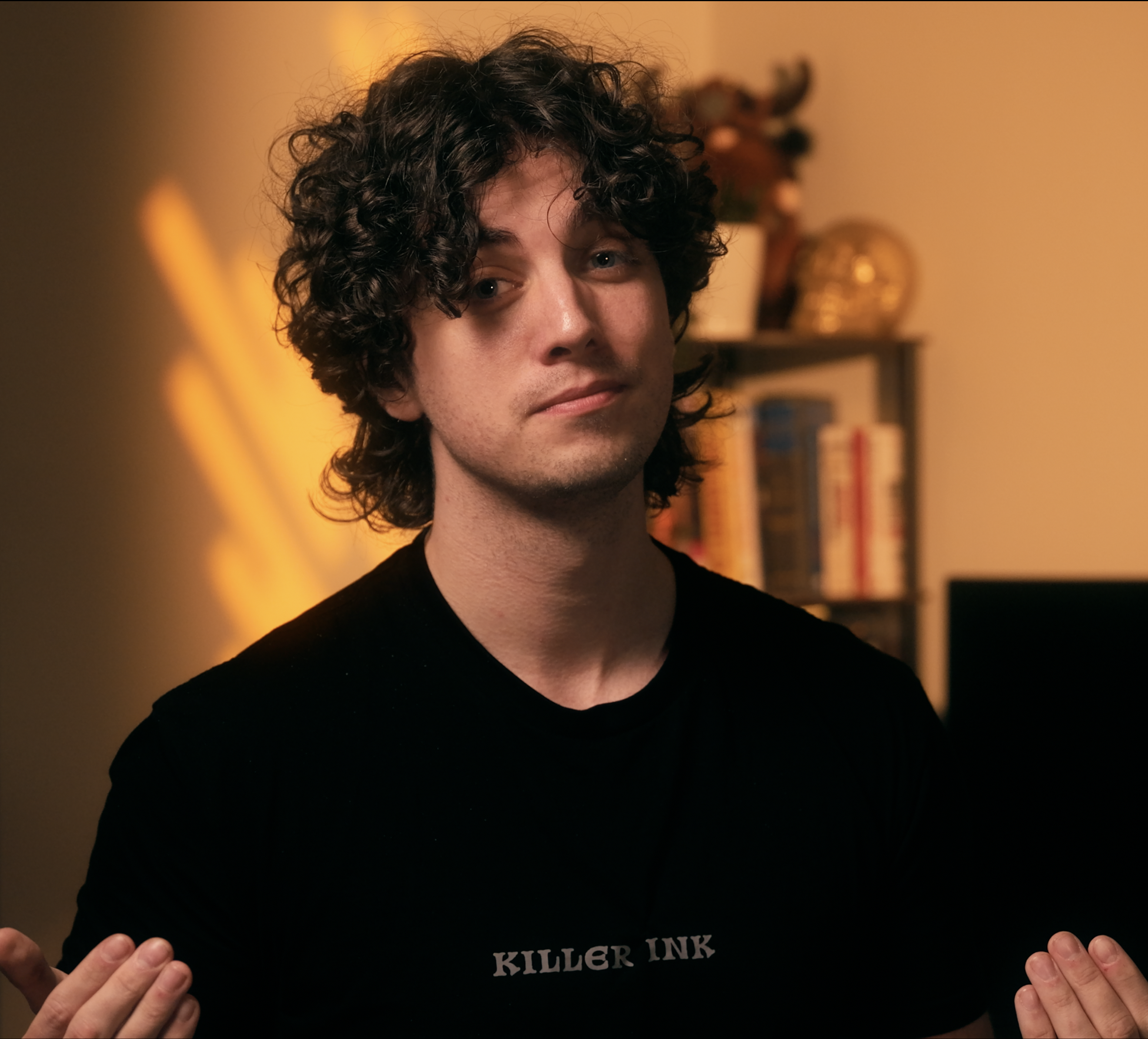A young man with curly dark hair and light skin wearing a black 'Killer Ink' t-shirt, sitting with hands raised, in a warmly lit room with a bookshelf and decorations in the background.