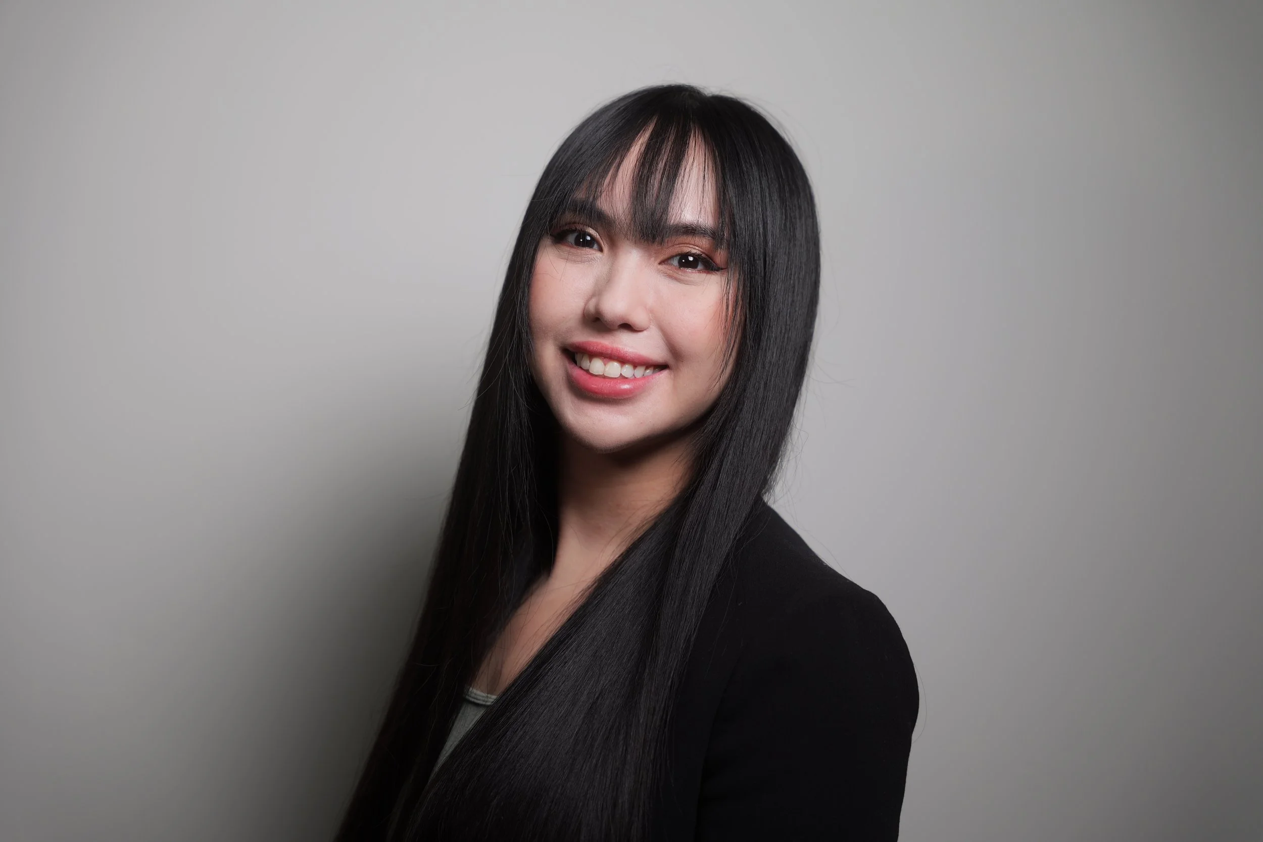 A young woman with long black hair, smiling, wearing a black blazer, standing against a plain gray background.