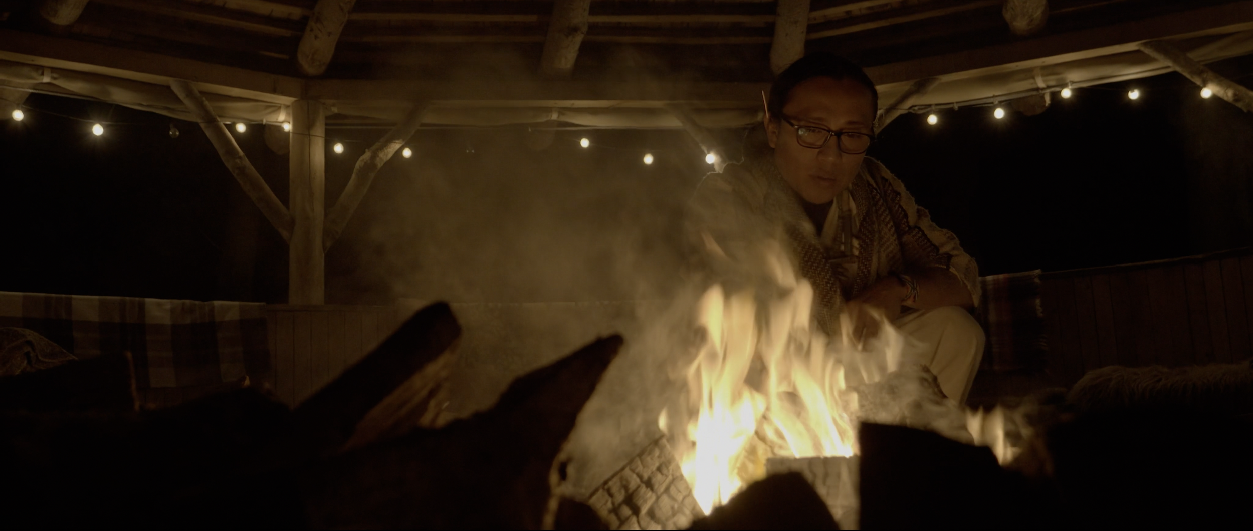 A man wearing glasses and a patterned shirt sits near a firepit outside at night under a wooden roof, with string lights overhead, and looks towards the fire.