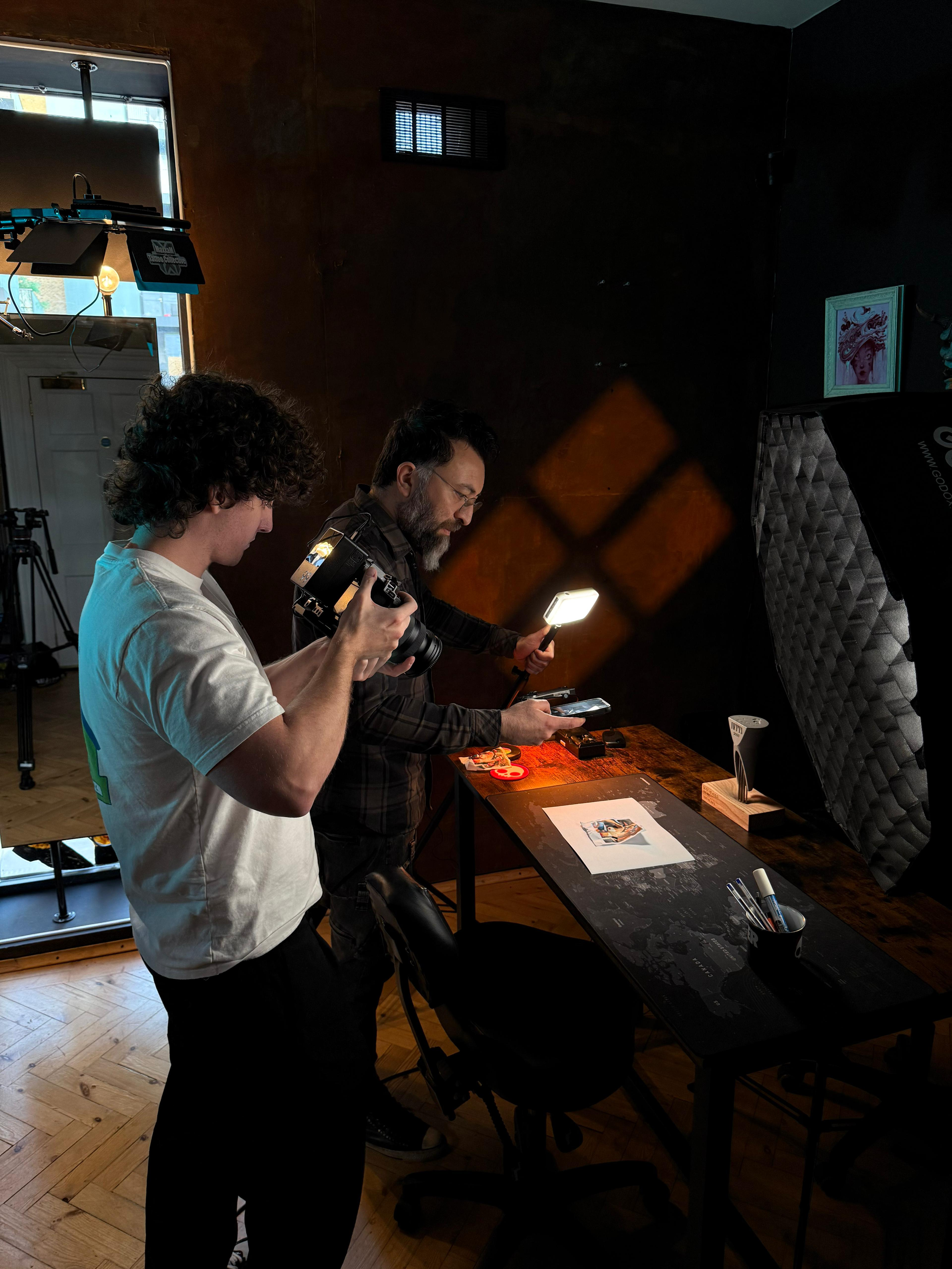 Two men are photographing an art piece on a desk, with professional lighting, studio equipment, and a dark background.