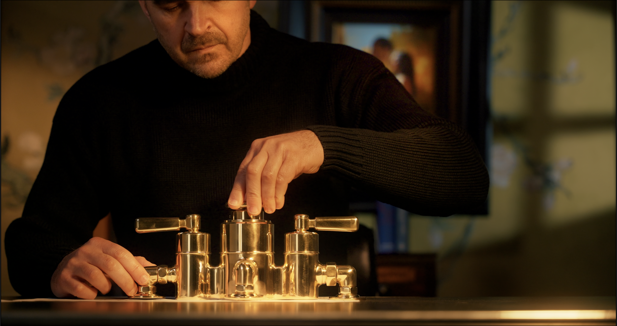 A man in a black sweater operating a shiny gold-colored mechanical device on a table in a warmly lit room.