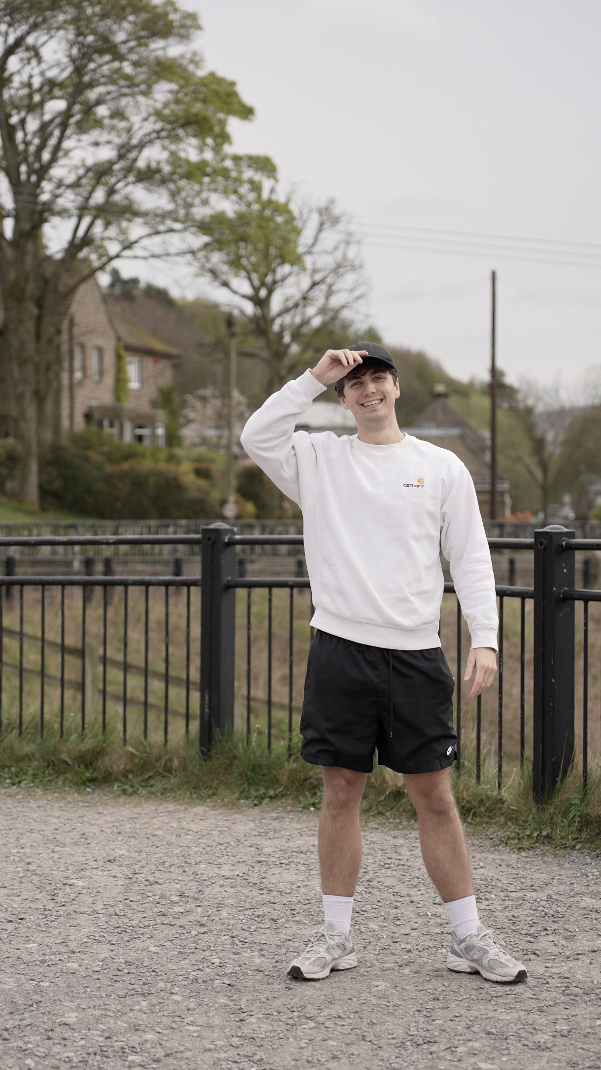 A young man smiling and tilting his cap, standing outdoors on a gravel path with a metal fence, trees, and houses in the background.