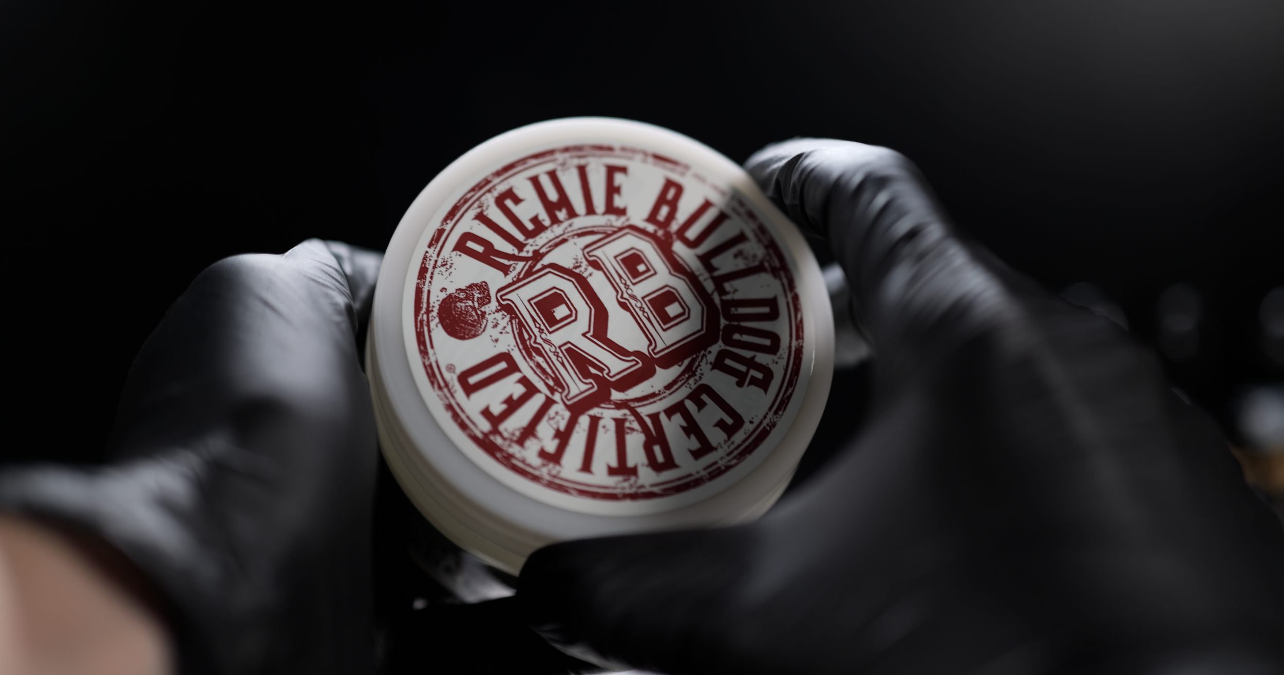 A hand wearing a black glove holds a circular item with a red and white logo, featuring the text "Richie Bully" and a heart graphic, on a black background.