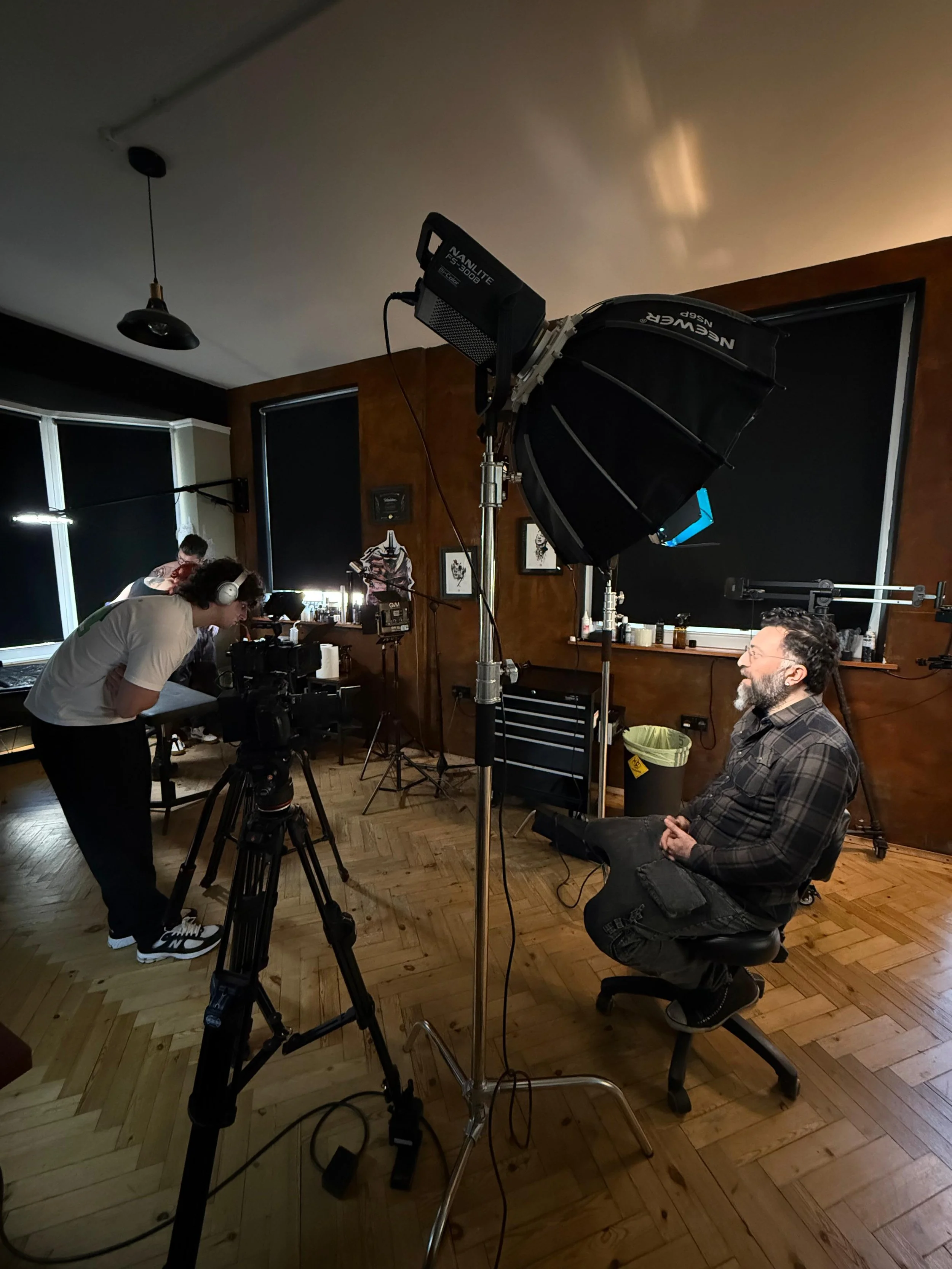 A man with glasses and a beard sitting on a chair during a video shoot, with a camera operator filming him, and professional lighting equipment set up in a room with wood-paneled walls and wooden floors.