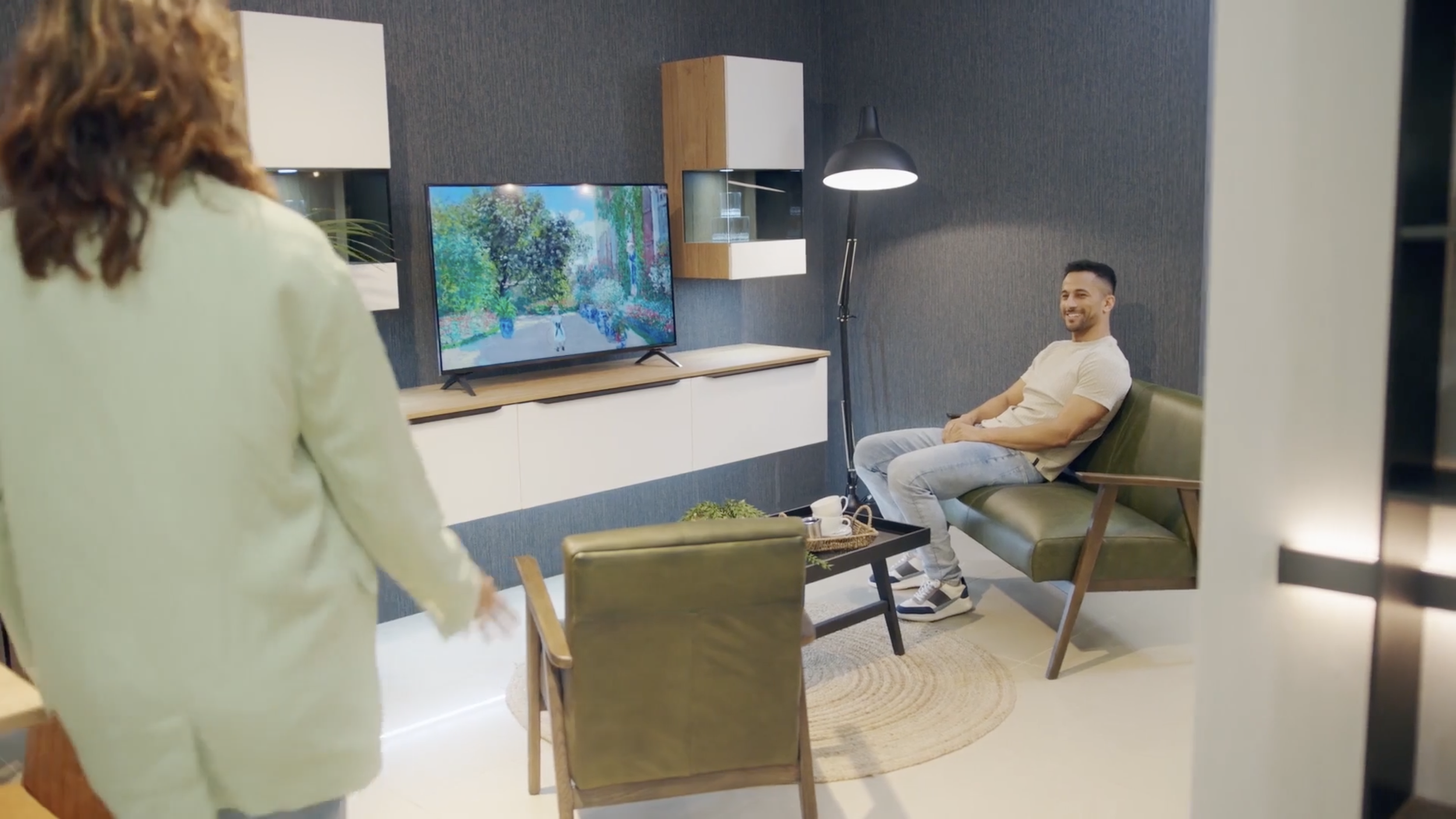 A man sits on a green sofa in a modern living room, smiling while watching TV. A woman stands nearby, partially visible. The room features dark textured walls, a flat-screen TV showing an outdoor scene, and contemporary furniture with a floor lamp and a table with cups.