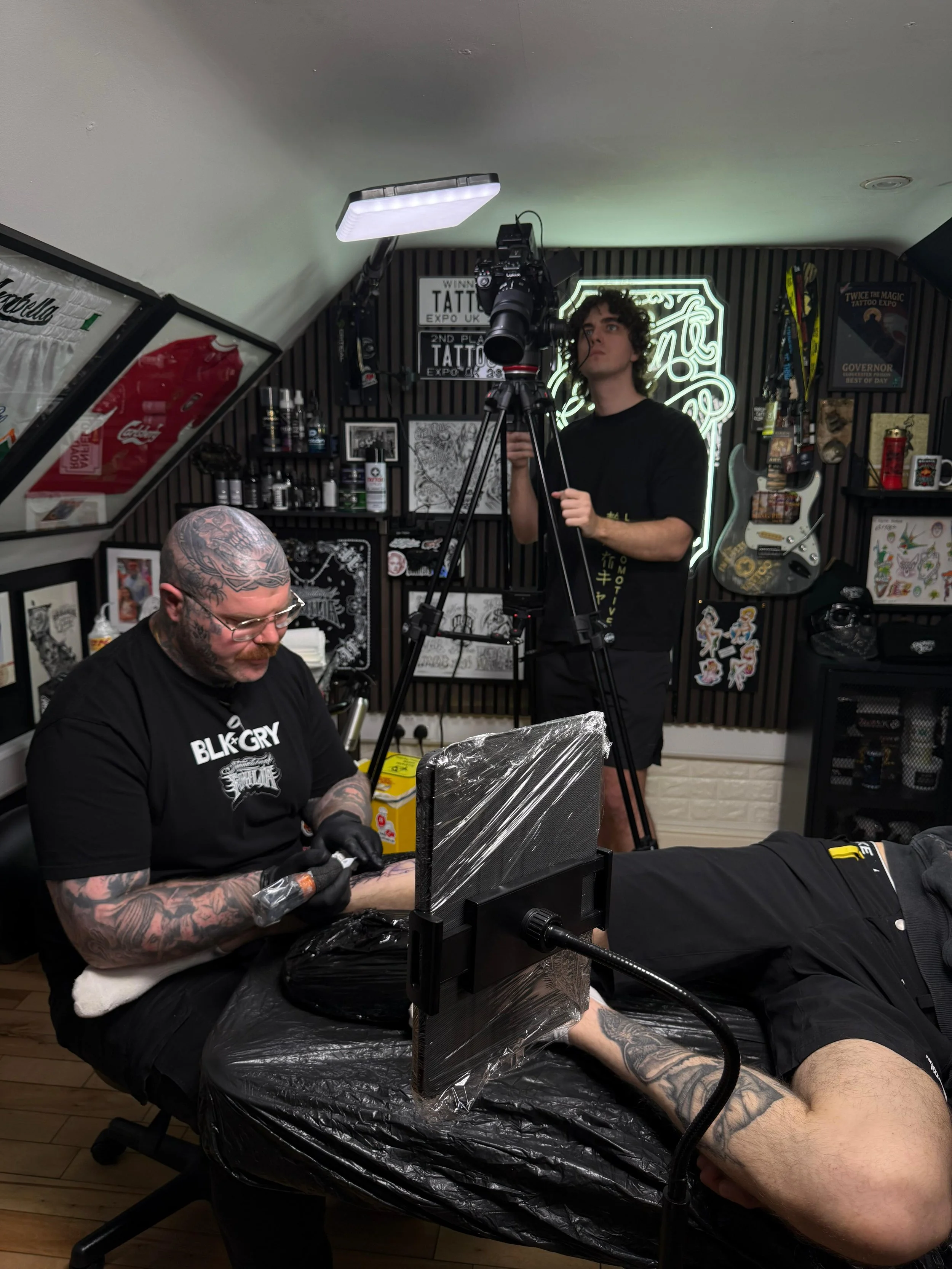 A tattoo artist with tattoos on his arms and head, wearing glasses and black gloves, is getting ready to tattoo a person's leg using a tattoo machine. The person on the table has light skin and is lying down with their leg exposed. The setting is a tattoo studio with various artwork, equipment, and posters on the walls, illuminated by a neon sign.