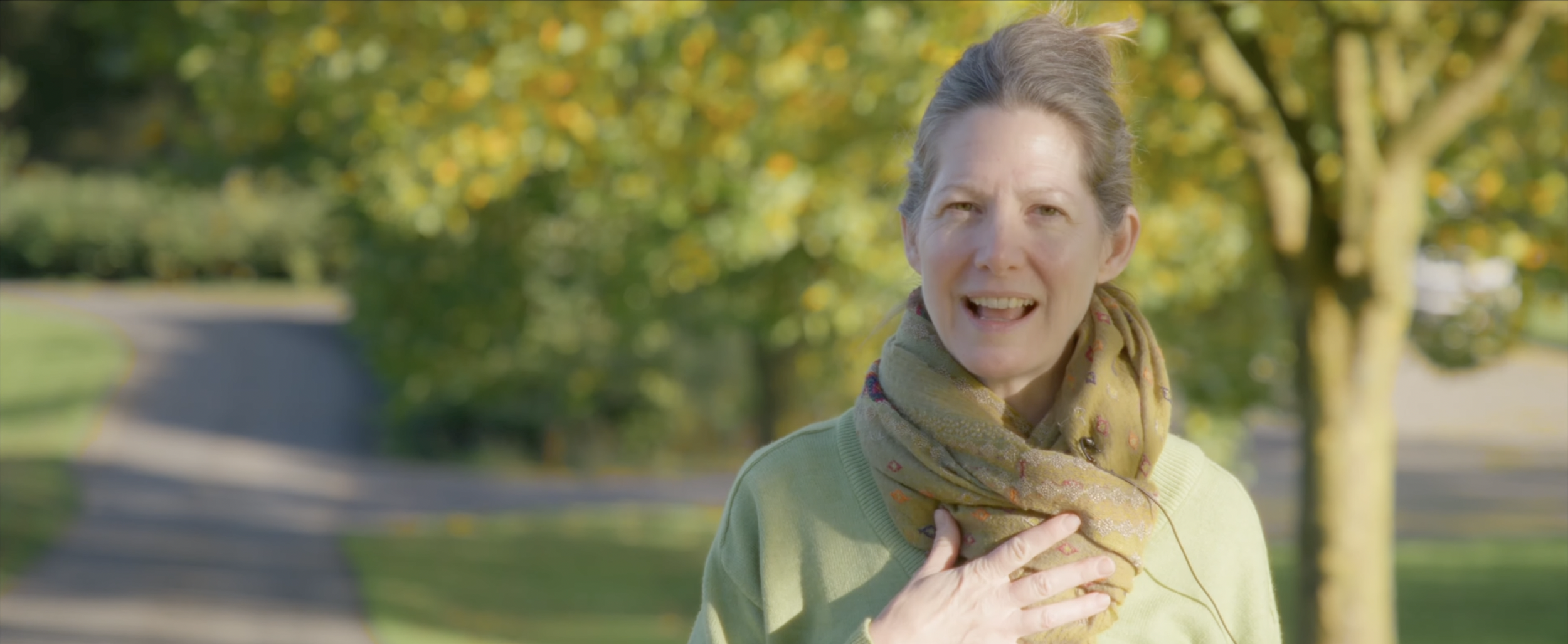 A woman with gray hair, wearing a green sweater and a patterned scarf, outdoors on a sunny day, speaking with her hand on her chest.