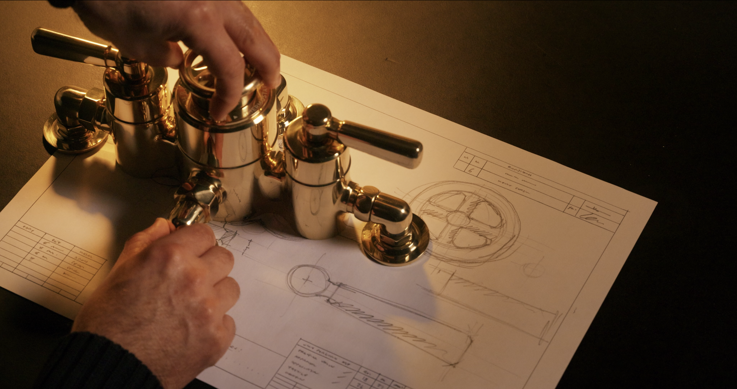 Hands working on a shiny metal faucet prototype placed on technical drawings and blueprints at a workspace.