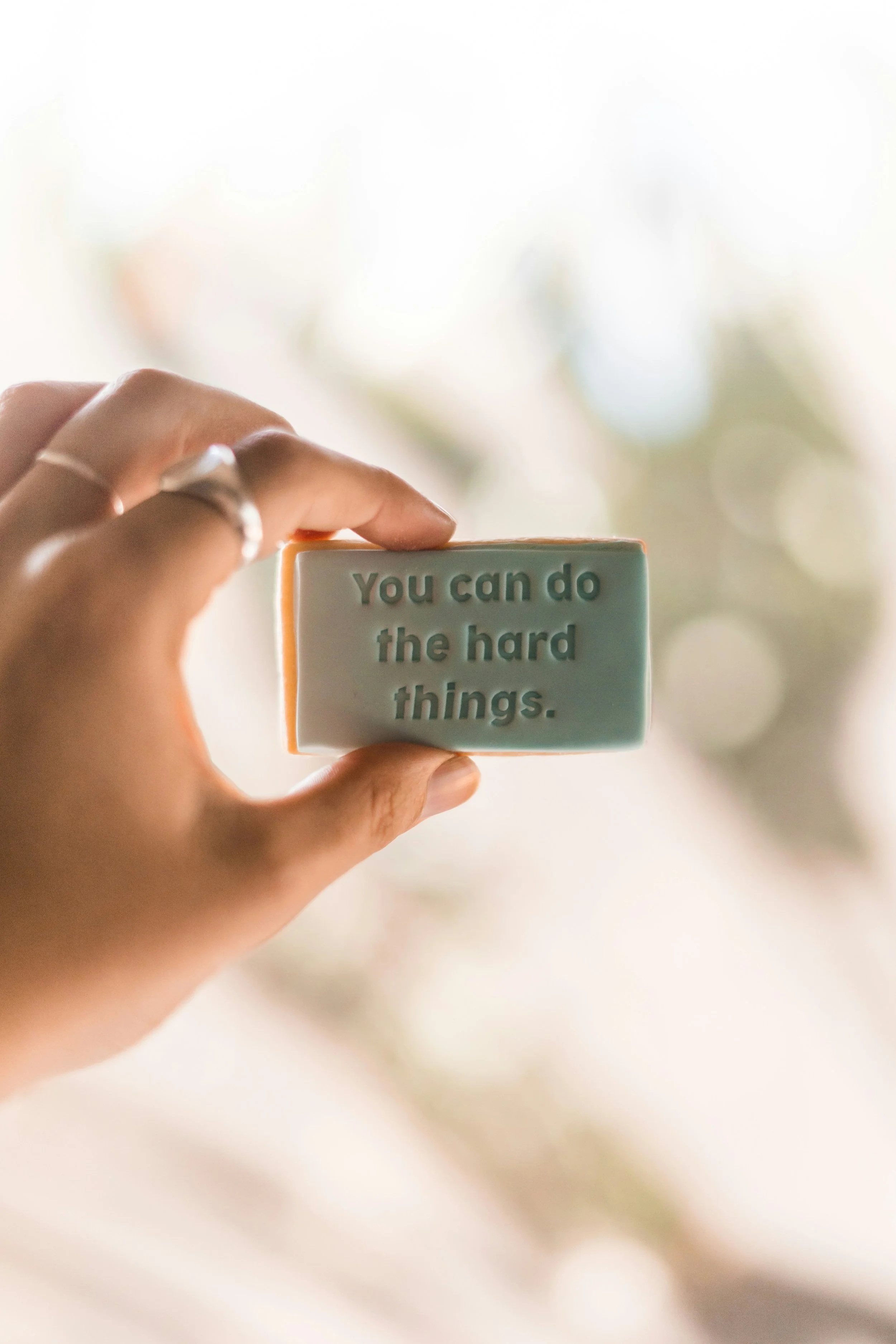 Close-up of a hand holding a rectangular soap with the words "You can do the hard things" inscribed on it, blurred background.