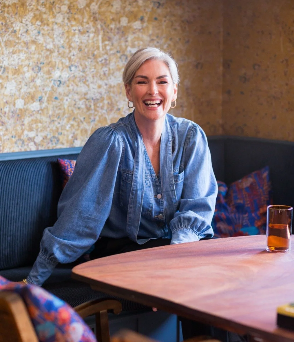 A woman with short gray hair and hoop earrings sitting at a table, smiling in a casual setting with colorful cushions and a textured wall in the background.