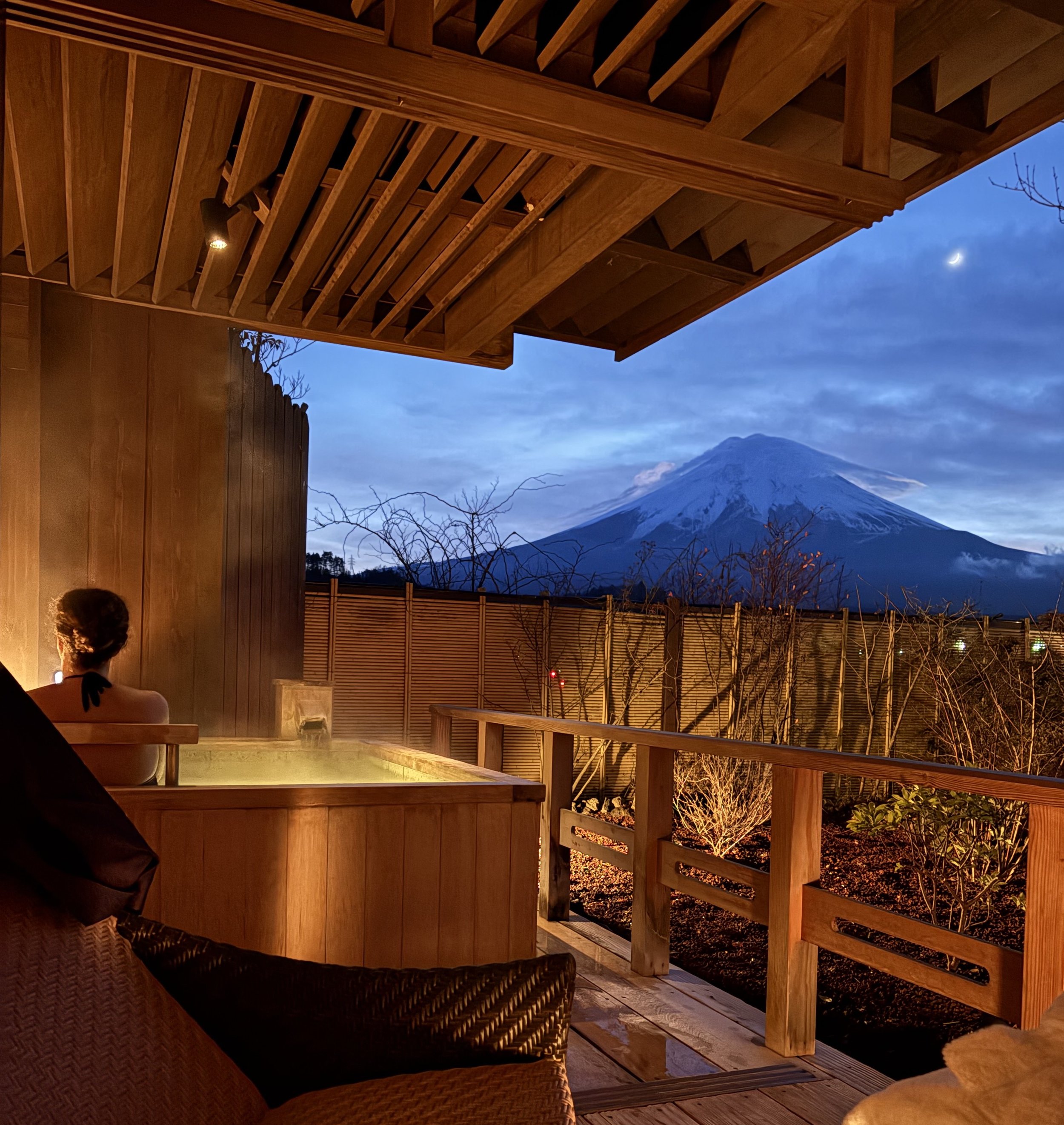 A person relaxing in an outdoor hot spring with a view of Mount Fuji at dusk, surrounded by wooden structures and fencing.