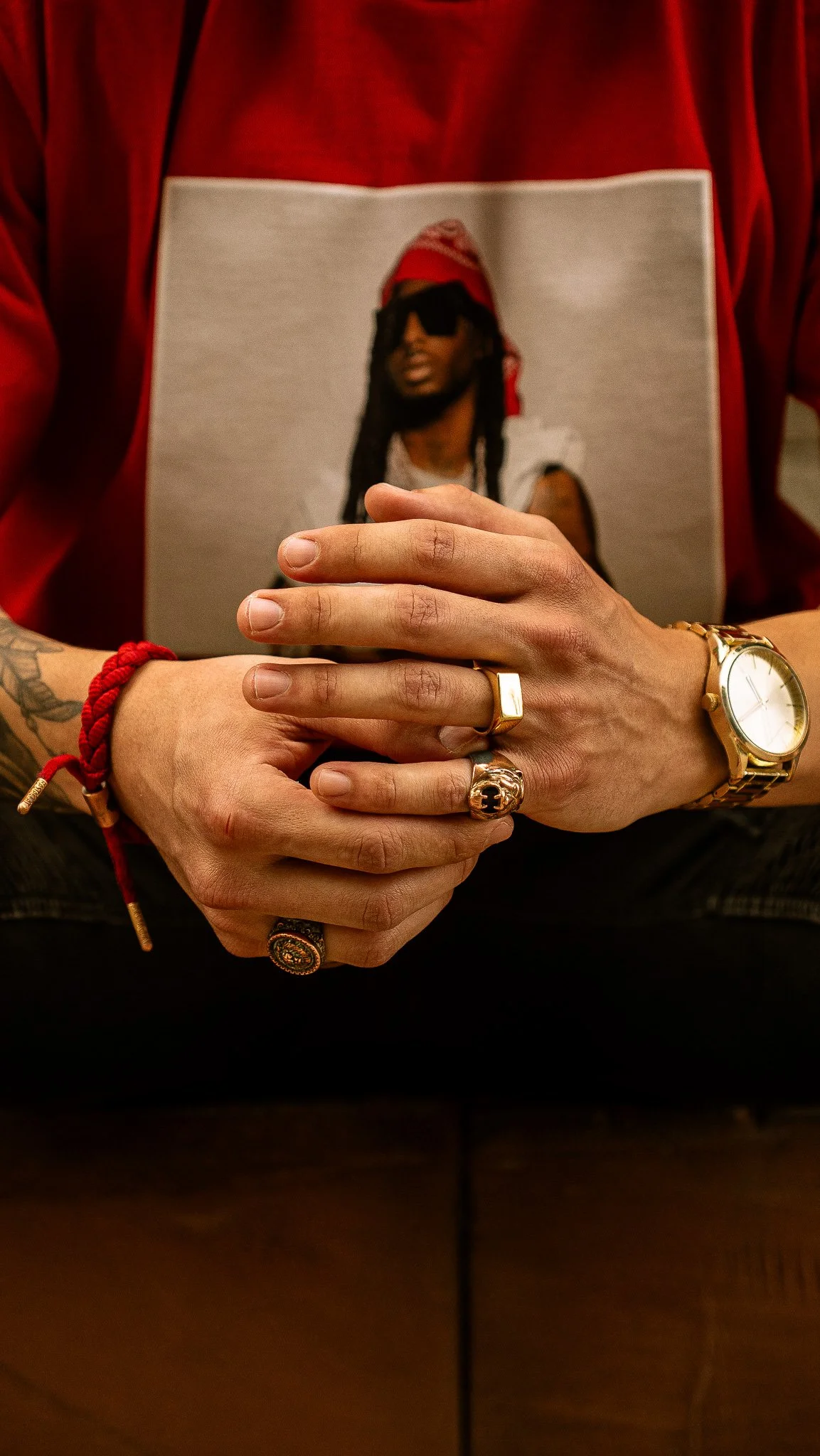 Person wearing a red shirt with a picture of a man with dreadlocks, sunglasses, and a red bandana, clasping their hands with various rings and watches.