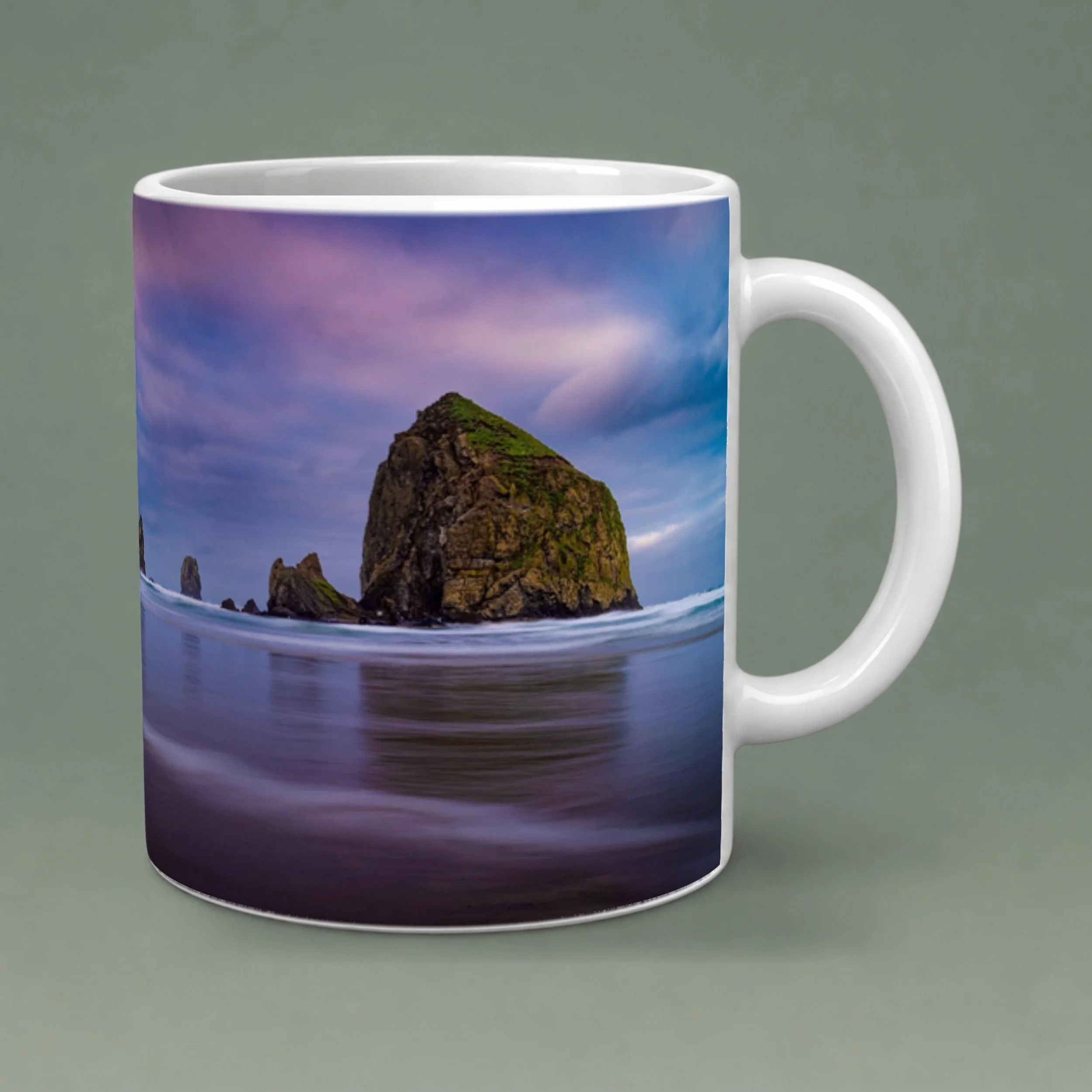 White 11 oz ceramic mug featuring Haystack Rock at Cannon Beach beneath a colorful sky, with a rounded handle and gray-green background.