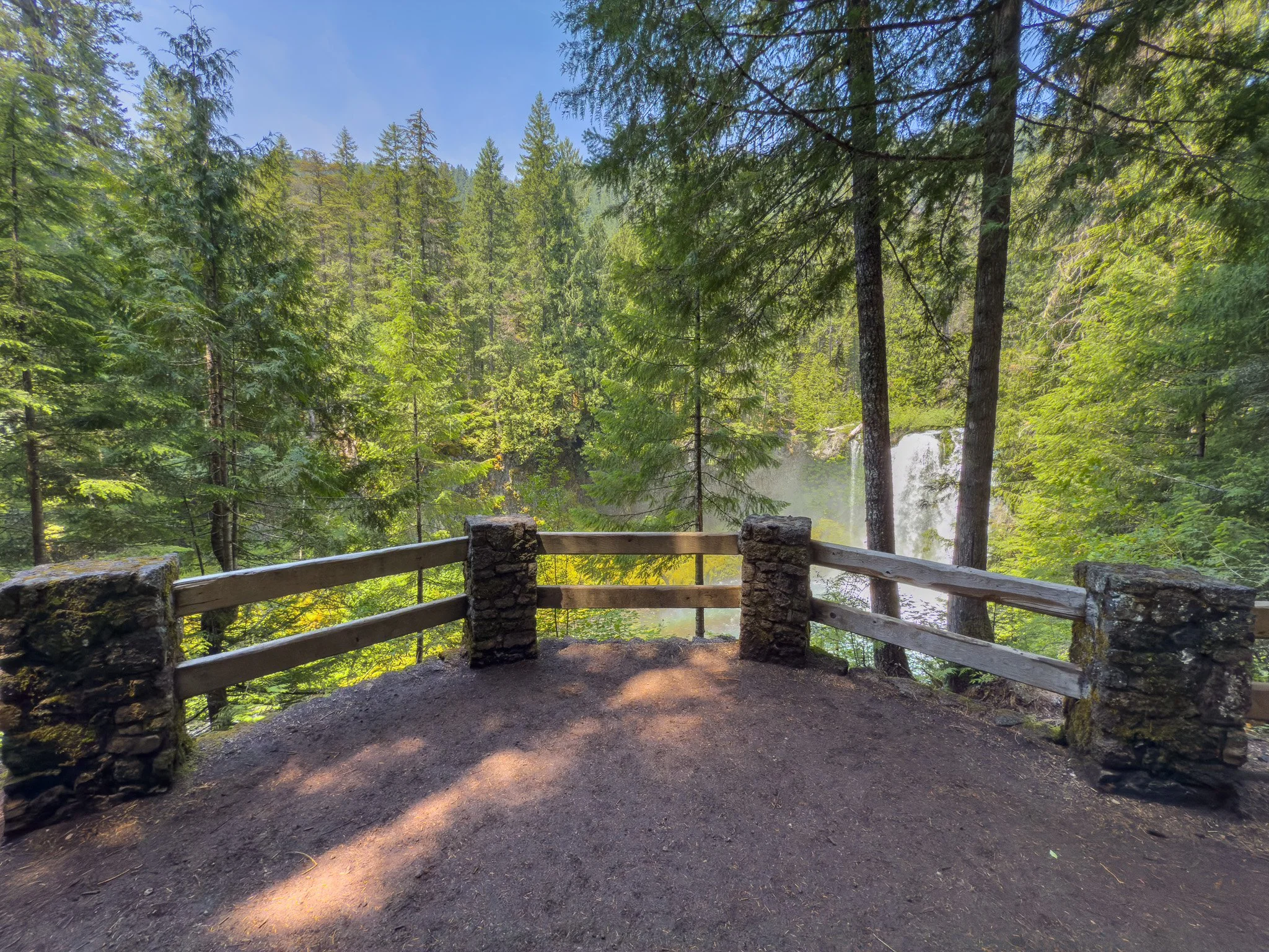 Main viewpoint overlooking Koosah Falls, framed by a rustic wooden and stone fence with tall evergreen trees surrounding the scene.
