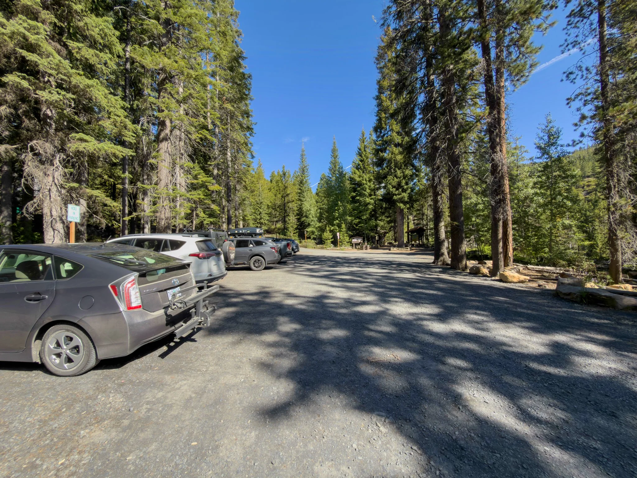 Gravel parking area at the Tumalo Falls Trailhead, with several parked vehicles surrounded by towering evergreen trees under a clear blue sky.