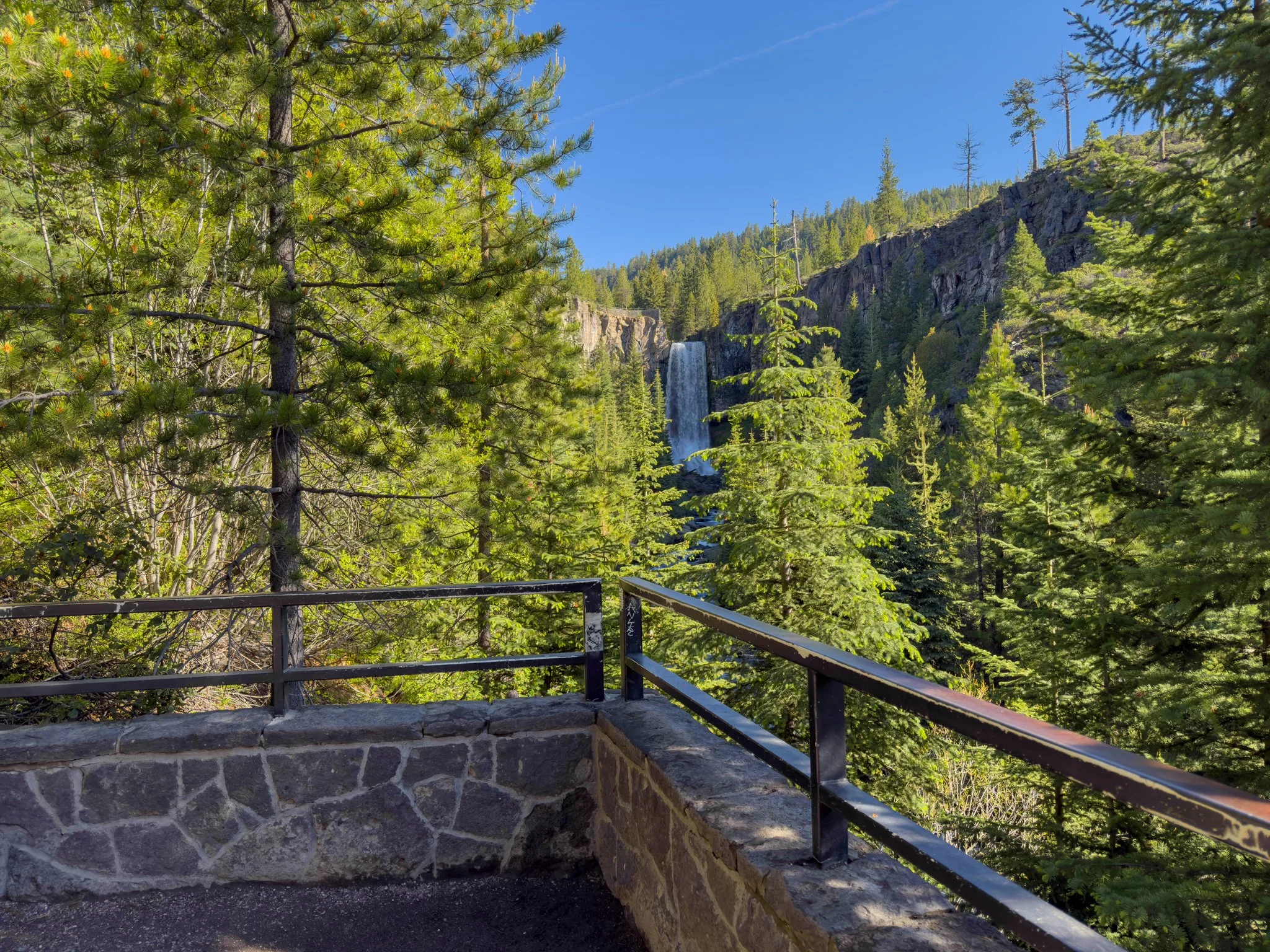 Main lower viewpoint overlooking Tumalo Falls, with the waterfall cascading over a tall basalt cliff and surrounded by evergreen trees beneath a clear blue sky.