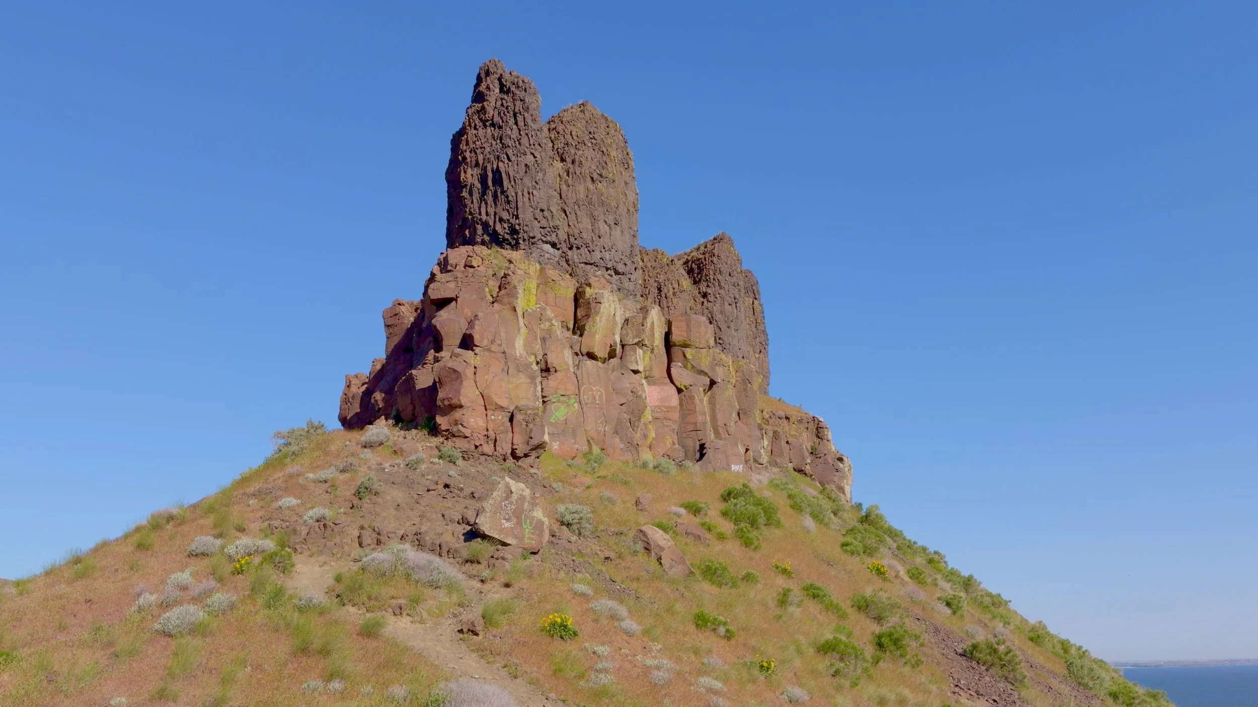> Close-up aerial view of Twin Sisters Rock's tall basalt columns rising dramatically from a rocky hilltop against a clear blue sky, with the Columbia River visible at the edge of the frame.