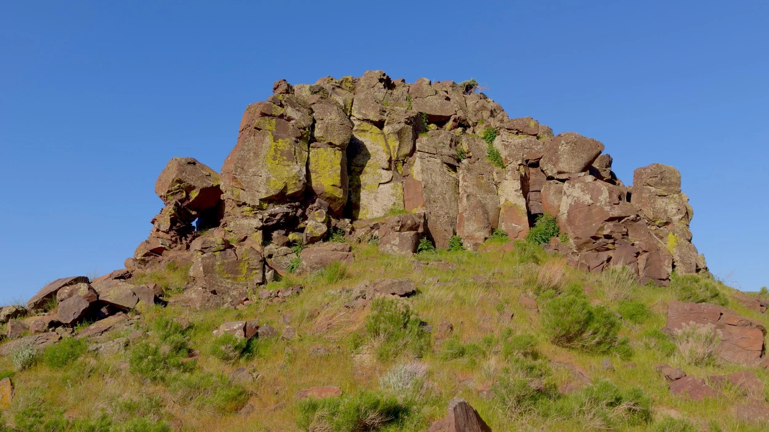 Close-up of a rugged basalt rock formation with lichen-covered columns rising above a green grassy hillside near Twin Sisters Rock, Washington.