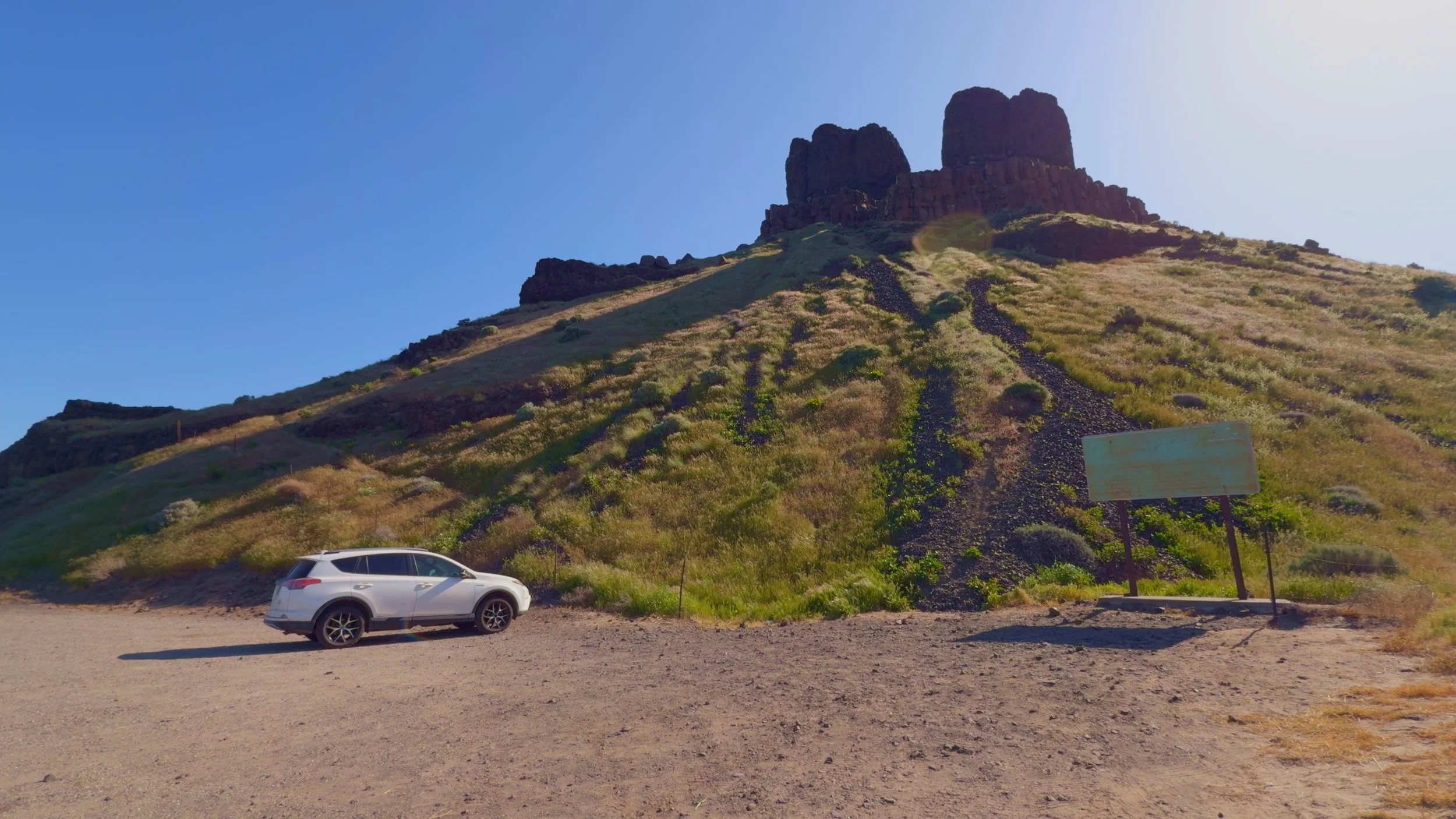 Gravel trailhead parking area at Twin Sisters Rock, Washington, with a white SUV and information sign below the basalt rock formation.