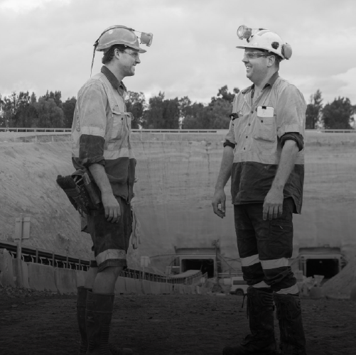 Two miners in safety gear talking at a construction site, wearing helmets with lights, work shirts, and protective boots, standing in front of an excavation pit.