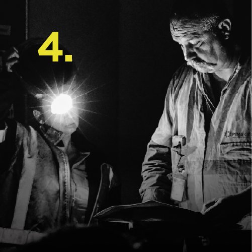 Two miners underground, one with a headlamp, examining documents in dim light.