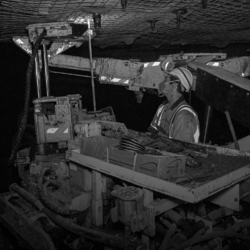 A miner operating machinery in an underground mine, equipped with a hard hat and safety gear, surrounded by equipment and tools.