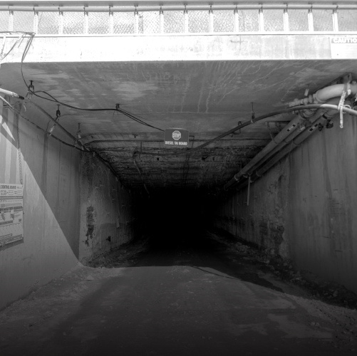 Concrete tunnel with exposed pipes and a partially visible warning sign, leading into darkness. The walls and ceiling show signs of wear and exposed wiring.