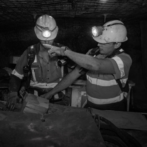 Two miners wearing hard hats with lights work underground, examining equipment in a dimly lit tunnel.