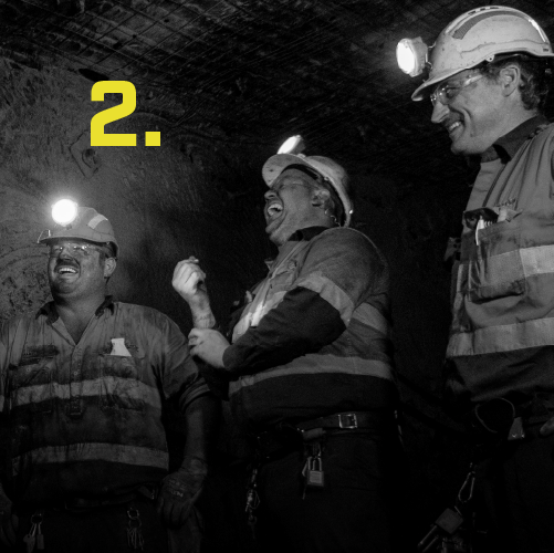 A group of miners wearing hard hats and headlamps standing in an underground tunnel, laughing and conversing. The number '2' is displayed in yellow on the left side of the image.