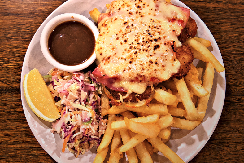 Plate of food with breaded fried chicken topped with melted cheese and ham, a side of French fries, coleslaw, lemon wedge, and a small cup of brown gravy, all on a wooden table.