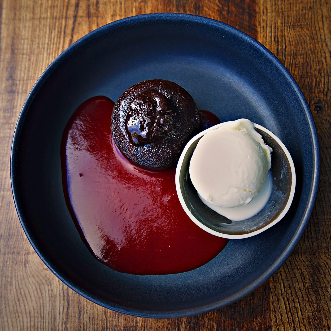 Chocolate lava cake with a scoop of vanilla ice cream, served on a dark blue plate with red sauce on a wooden table.