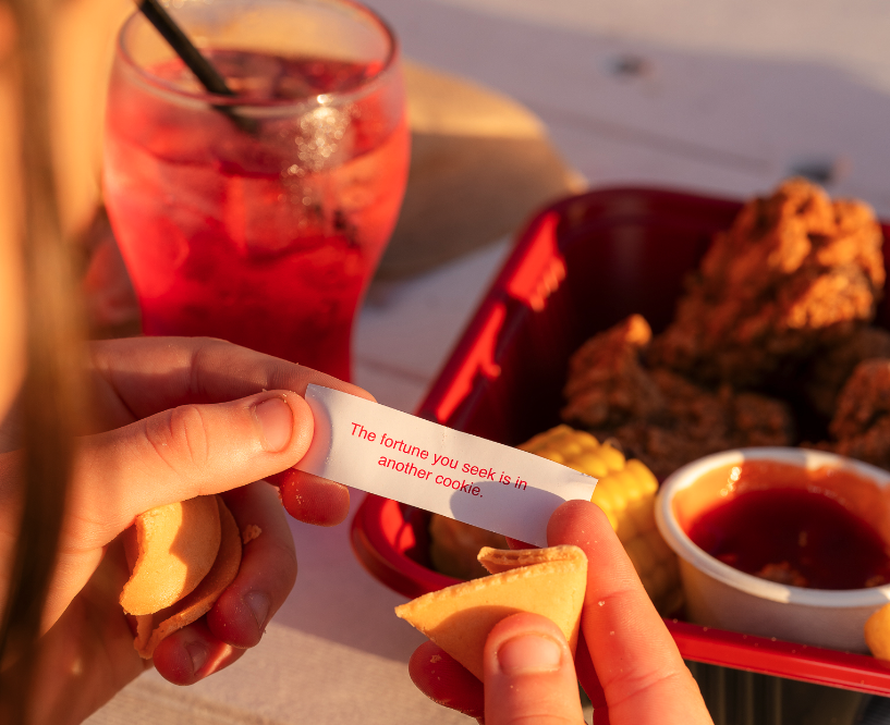 Person holding a fortune cookie with a paper reading 'The fortune you seek is in another cookie.' near a tray of fried chicken, corn on the cob, a cup of sauce, and a glass of pink drink on a table outdoors.