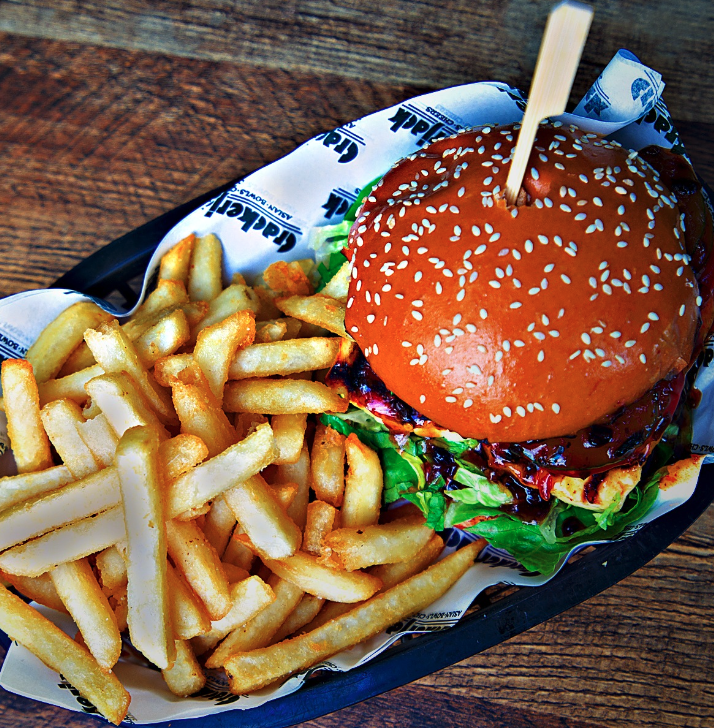 A basket containing a cheeseburger with lettuce, tomato, and barbecue sauce, topped with a sesame seed bun, accompanied by a side of French fries on a wooden table.