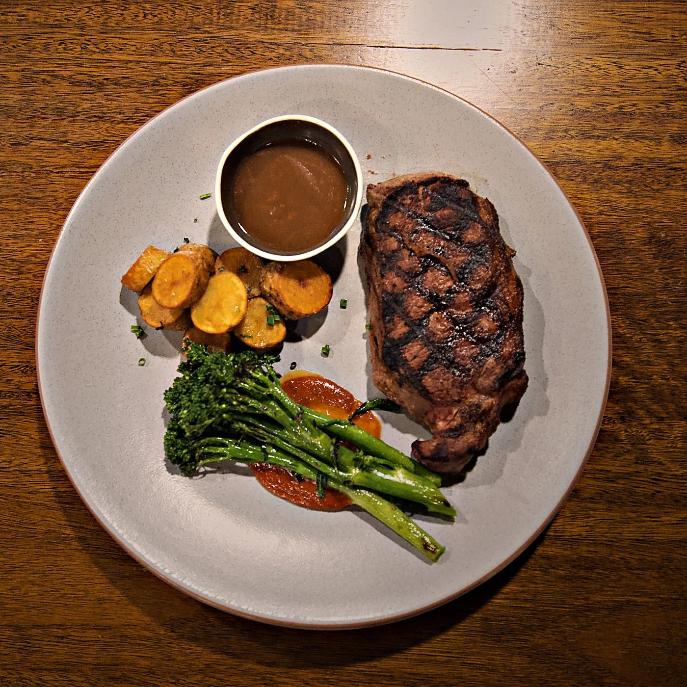 A plate with grilled steak, roasted sweet potatoes, broccoli, and a small cup of gravy on a wooden table.