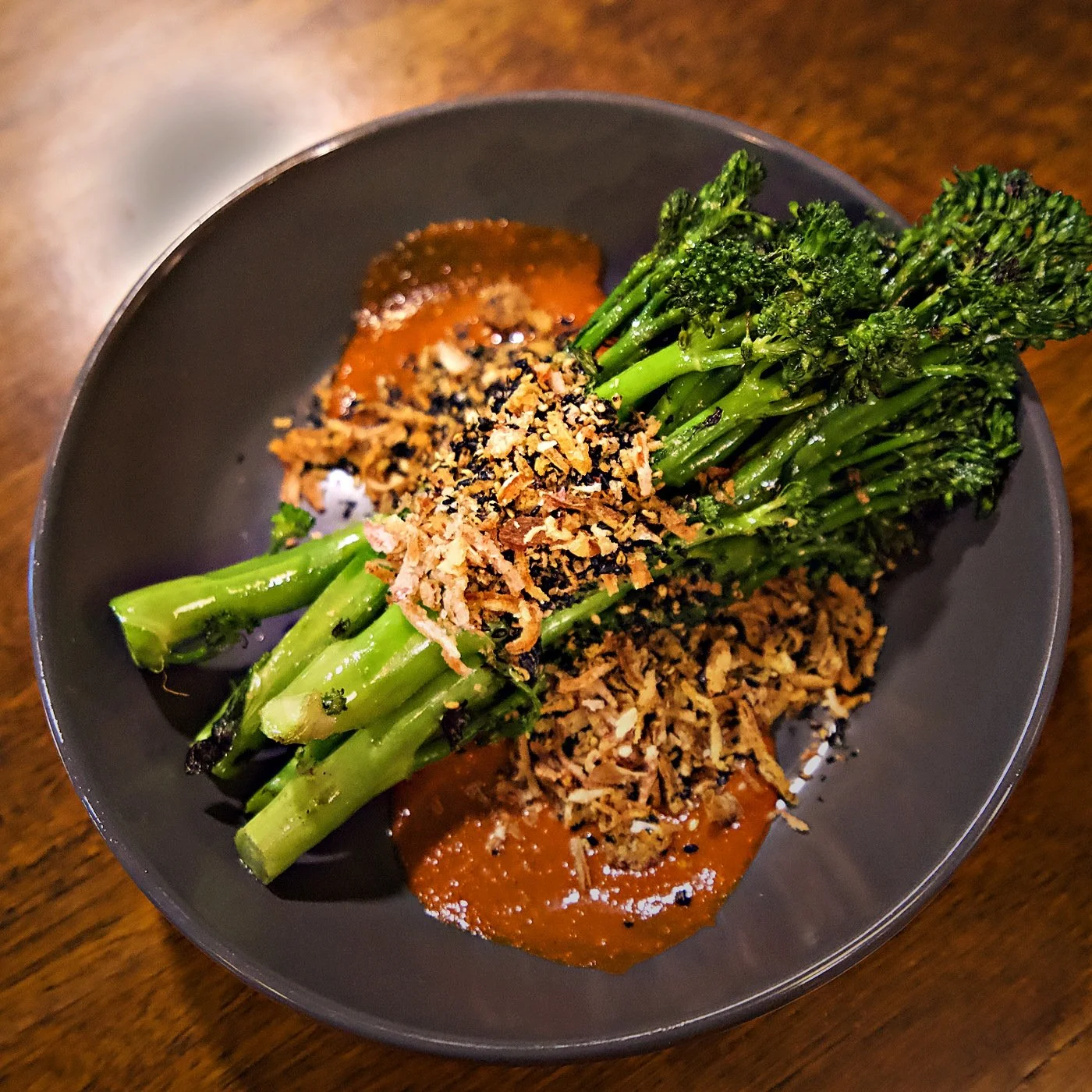 Plate of cooked vegetables including broccolini, topped with crispy onions, and drizzled with a brown sauce, on a dark plate placed on a wooden table.