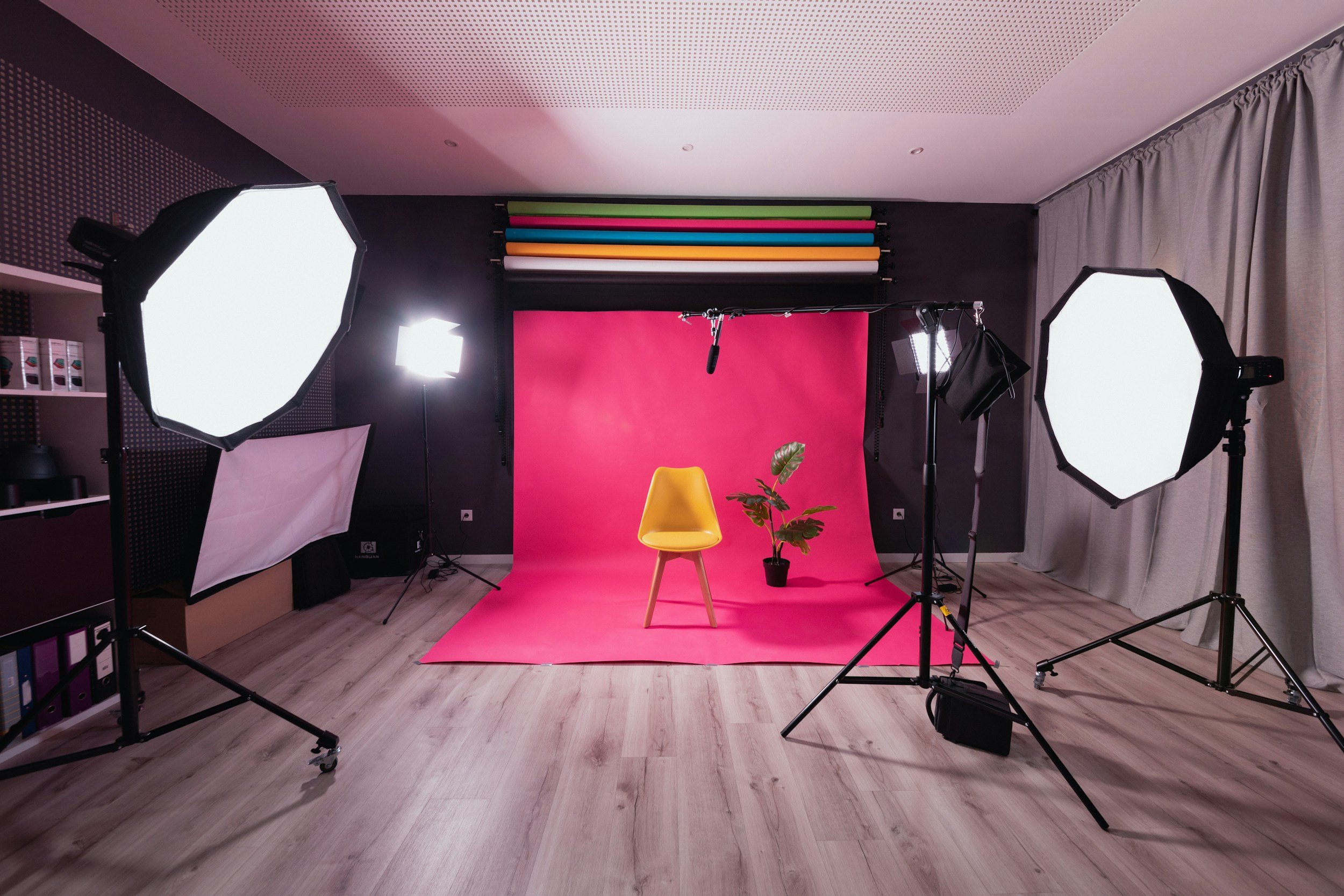 Estudio de fotografía con fondo rosa, sillón amarillo y planta en maceta, con iluminación profesional y equipo de grabación.