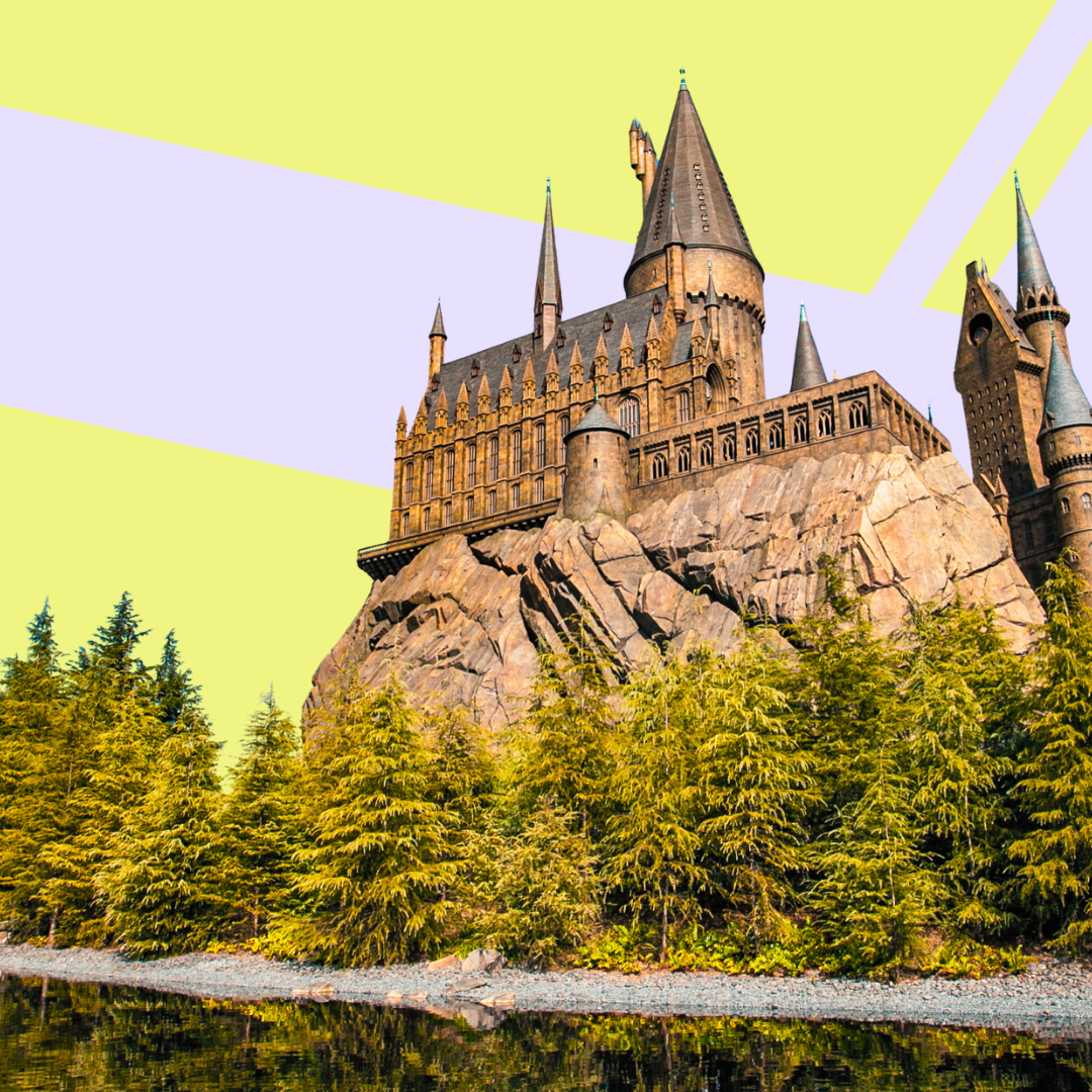 A castle with multiple towers and spires on a rocky hilltop, surrounded by green trees and reflected in a body of water.