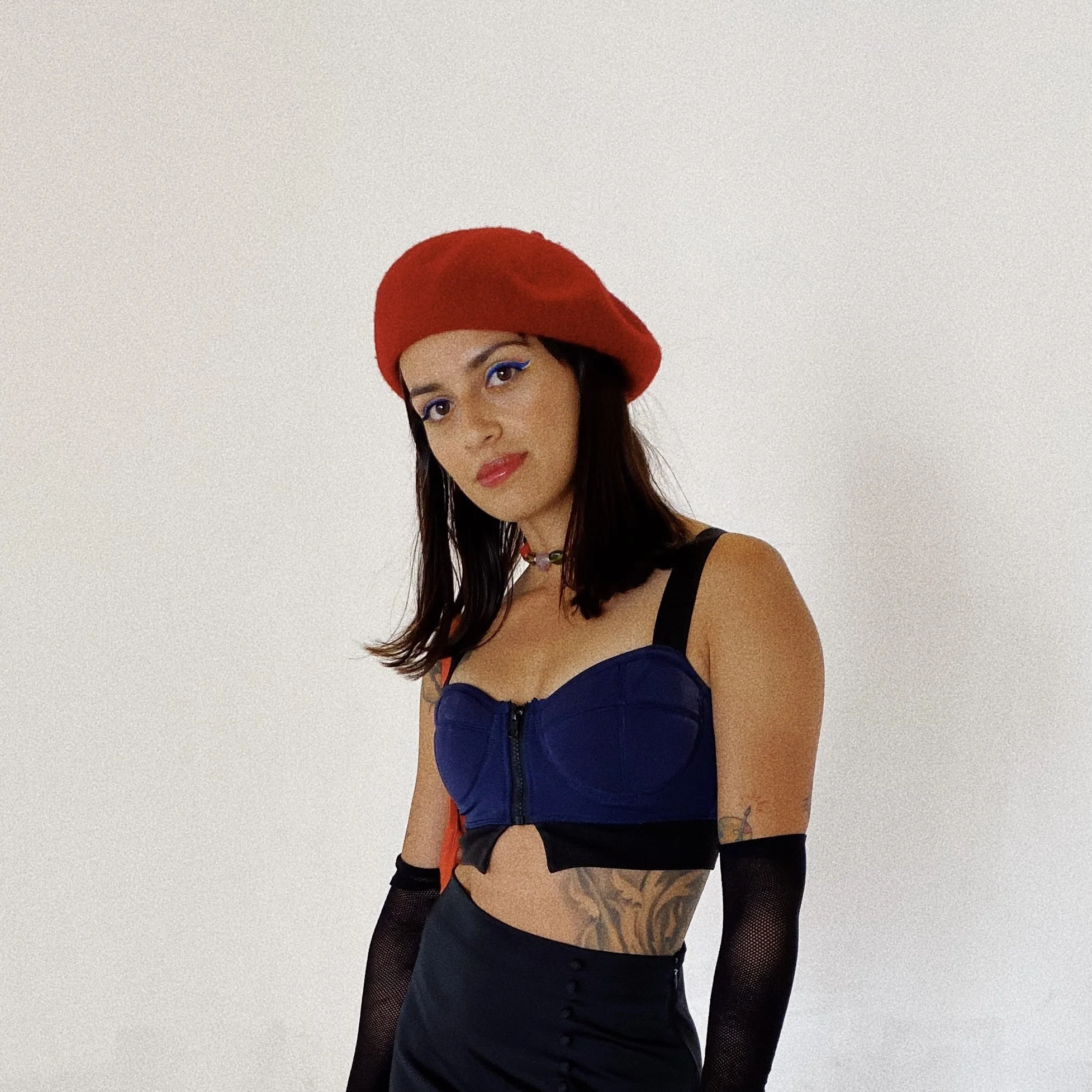 Woman with dark hair wearing a red beret, blue bustier, black skirt, and long black mesh gloves, standing against a plain white background.