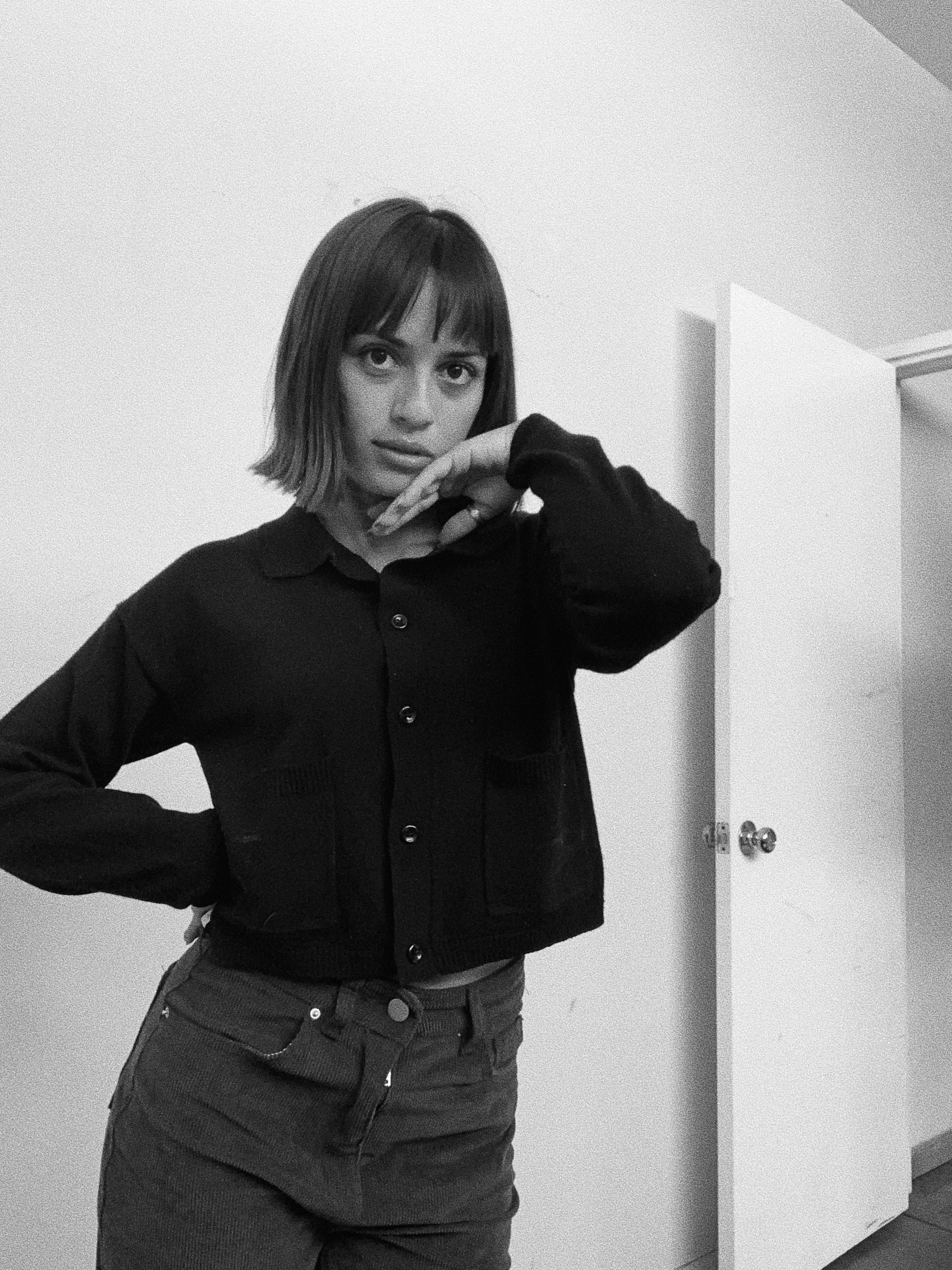 A woman with a bob haircut posing indoors in black and white, wearing a black jacket and dark pants, with one hand on her hip and the other near her face.