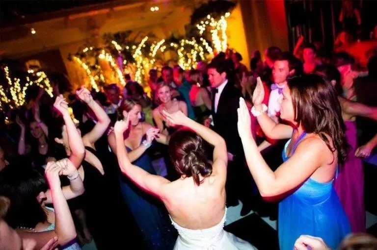 People dancing and enjoying themselves at a lively party or wedding reception with string lights and festive decorations.