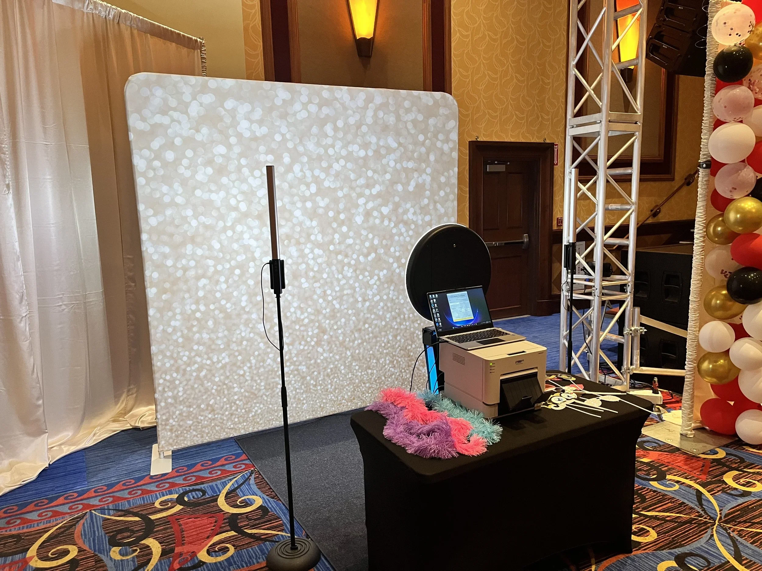 Photo of a photo booth setup with a white sparkly backdrop, a microphone stand, a laptop, a printer, and a colorful feather boa on a table. Part of a balloon column with red, white, black, and gold balloons and a stage-like area with metal trusses.