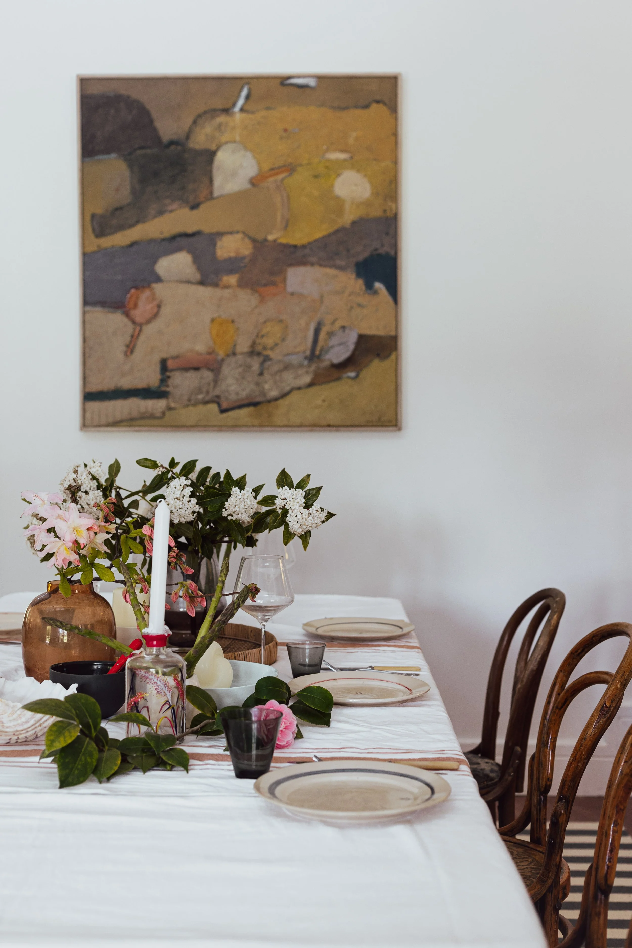A dining table decorated with flowers, plates, glasses, and candles, set for a meal in a room with a white wall and abstract art hanging on the wall.