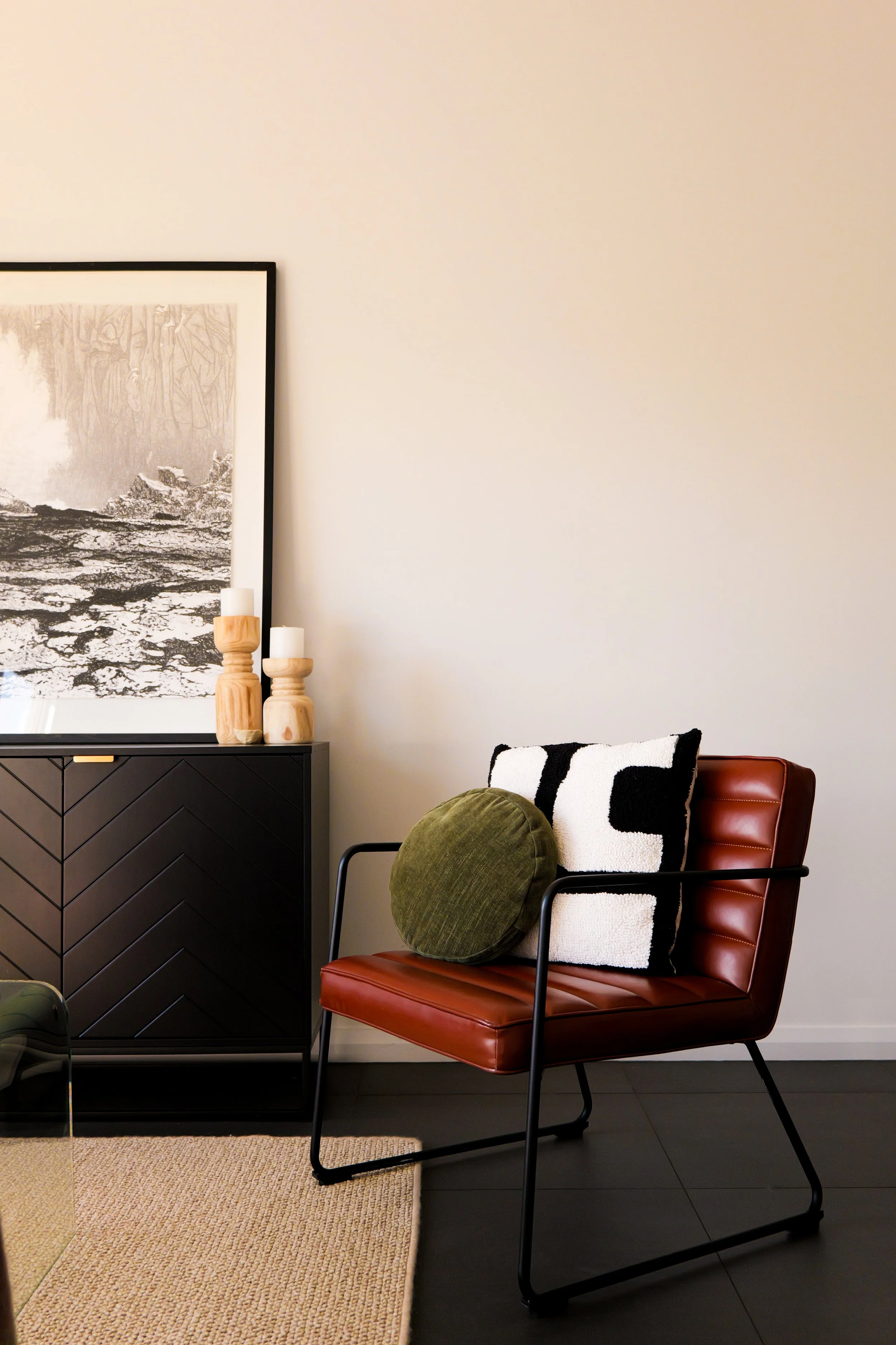 A modern living room corner with a brown leather chair, black metal frame, decorative pillows, and a black sideboard with artwork and candle decor.