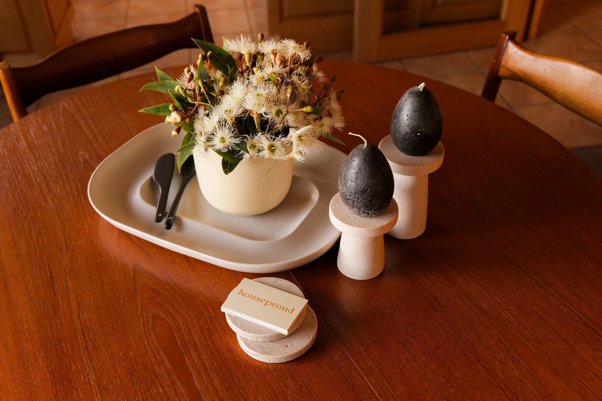Decorative table centerpiece with a white ceramic pot holding a bouquet of white and pink flowers with green leaves, placed on a white tray with black tongs, flanked by two black candles on stone candle holders, and a small stack of notes with the word 'houseproud' on top, all on a wooden table.