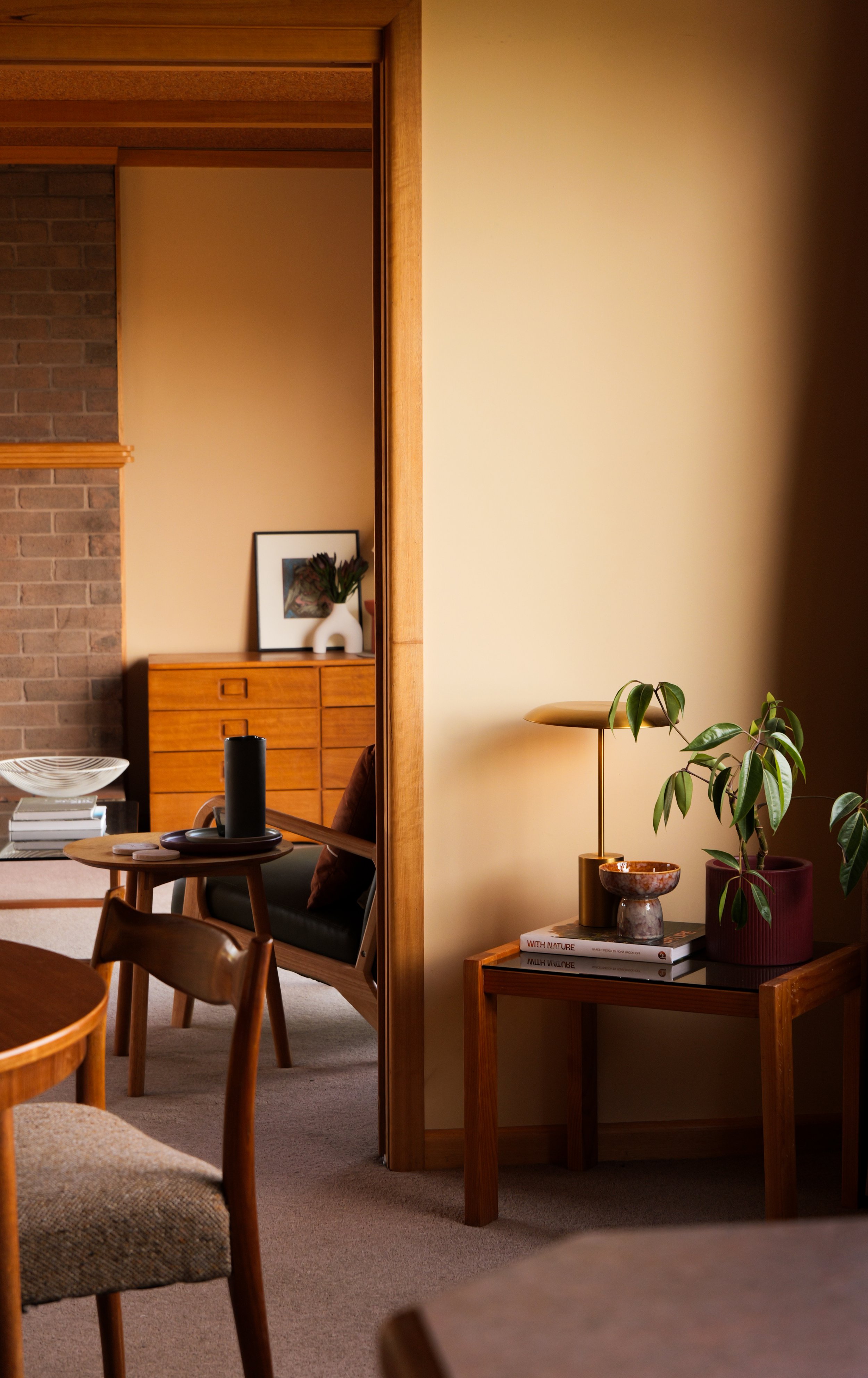 A cozy interior corner of a living room with a wooden side table holding a potted plant, book, and lamp, with a partial view of a sitting area and decor in the background.