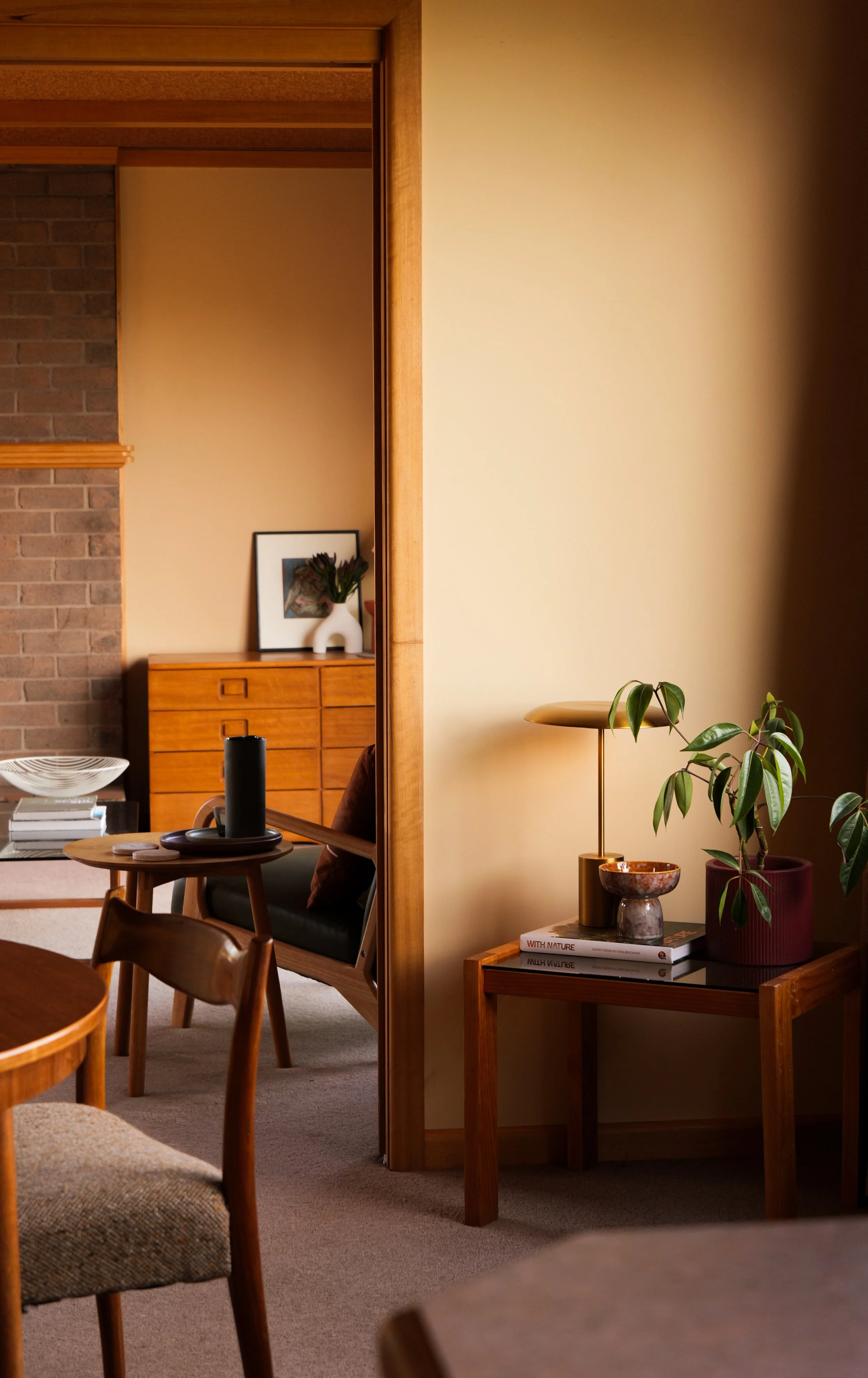 Interior view of a cozy living room with warm lighting, featuring wooden furniture, a green potted plant on a side table, and a background with a brick wall, framed artwork, and storage drawers.