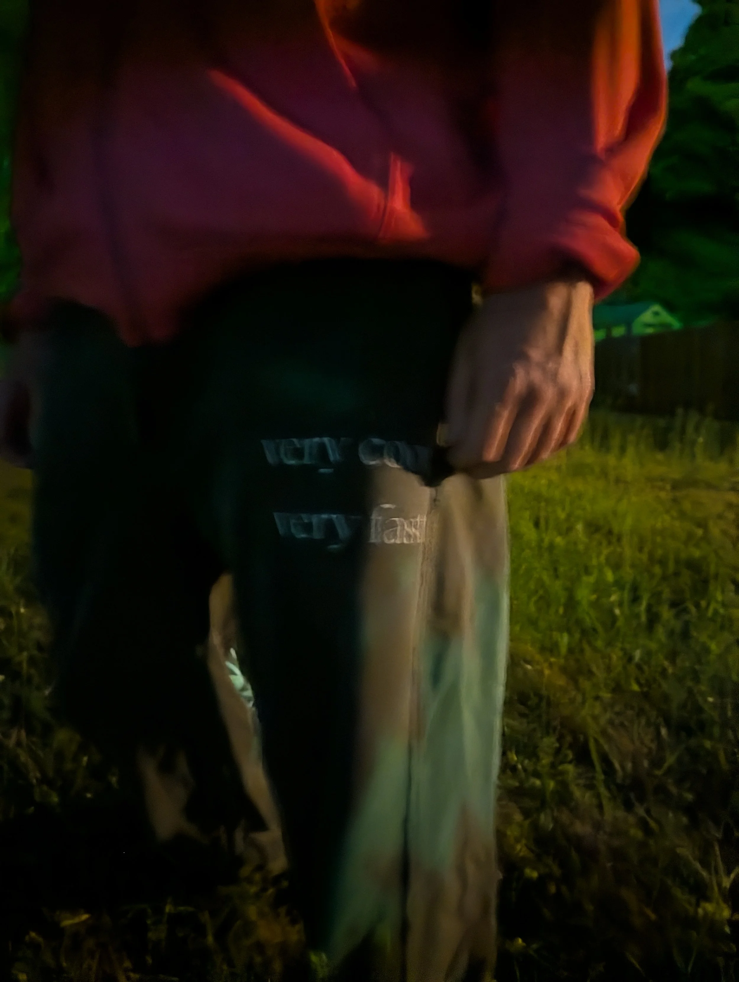 A person wearing a red jacket and black pants sitting on a wooden bench in a grassy outdoor area during nighttime, with a dark background and some greenery.