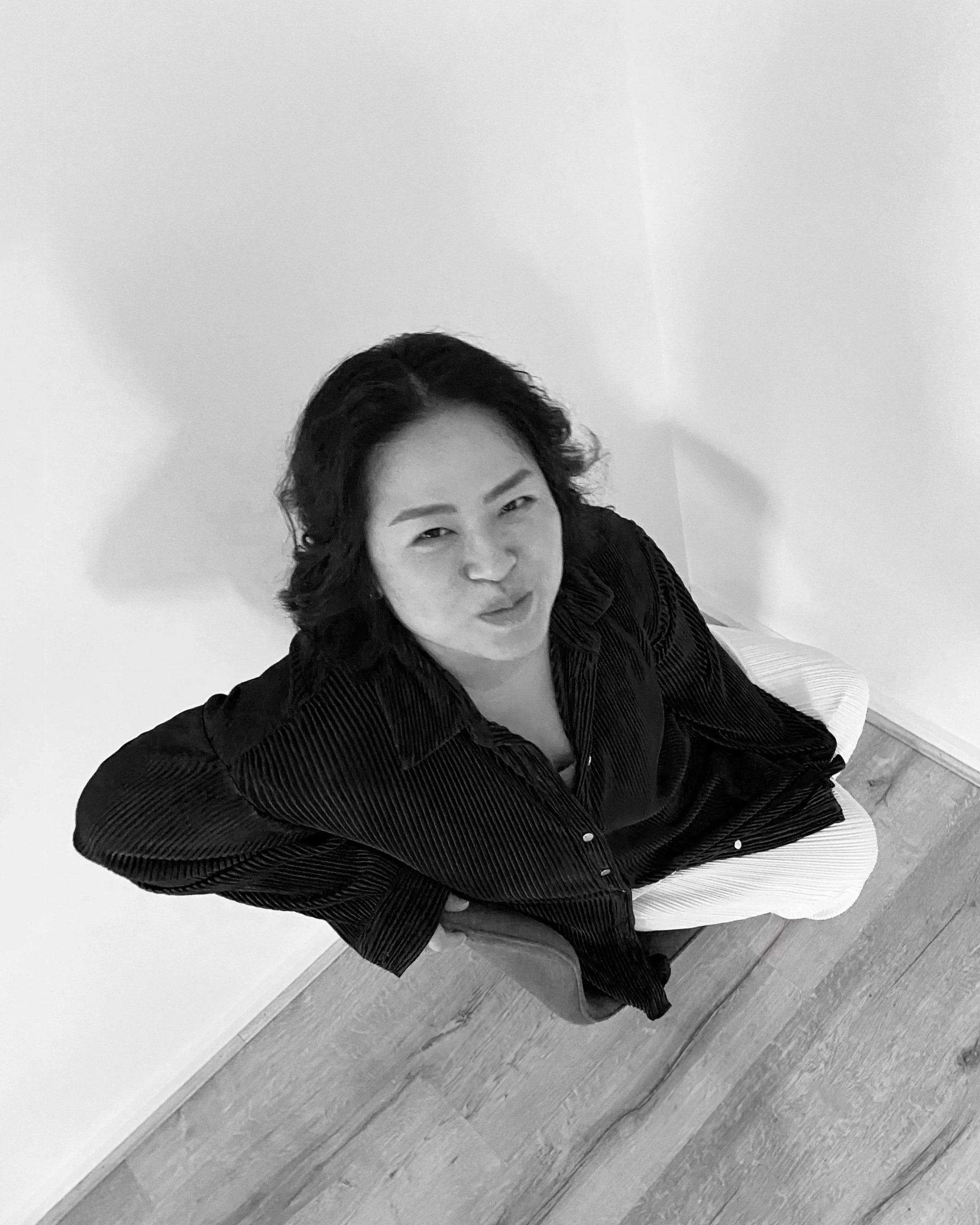 Nina with short curly hair wearing a dark striped shirt, smiling, seated on a white cushion on a wooden floor, against a plain white wall.