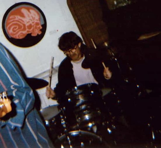 A person playing drums, sitting behind a drum set in a room with a white wall and a circular red dragon artwork.