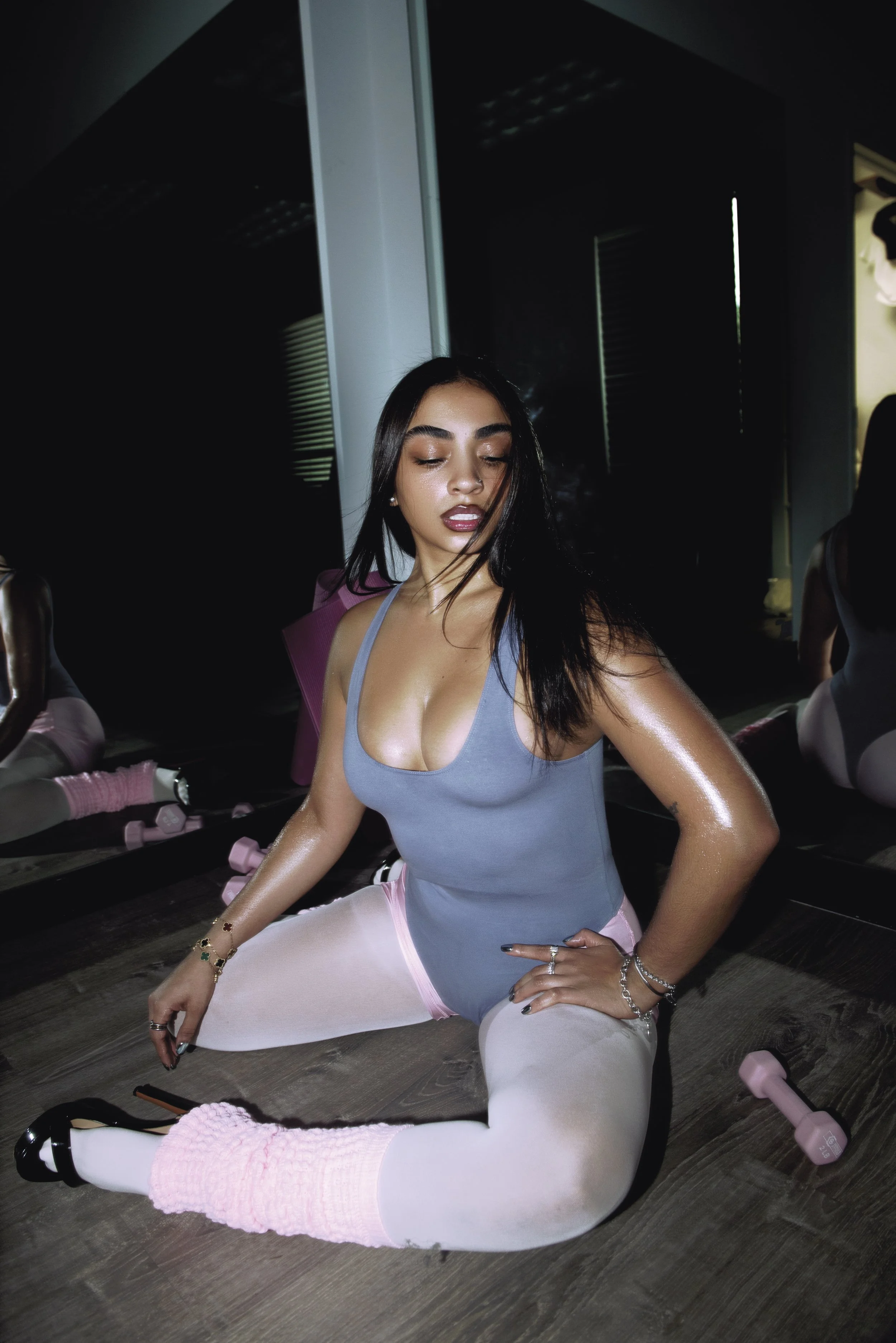 Woman in a blue tank top and pink pants kneeling on a gym floor beside pink dumbbells.